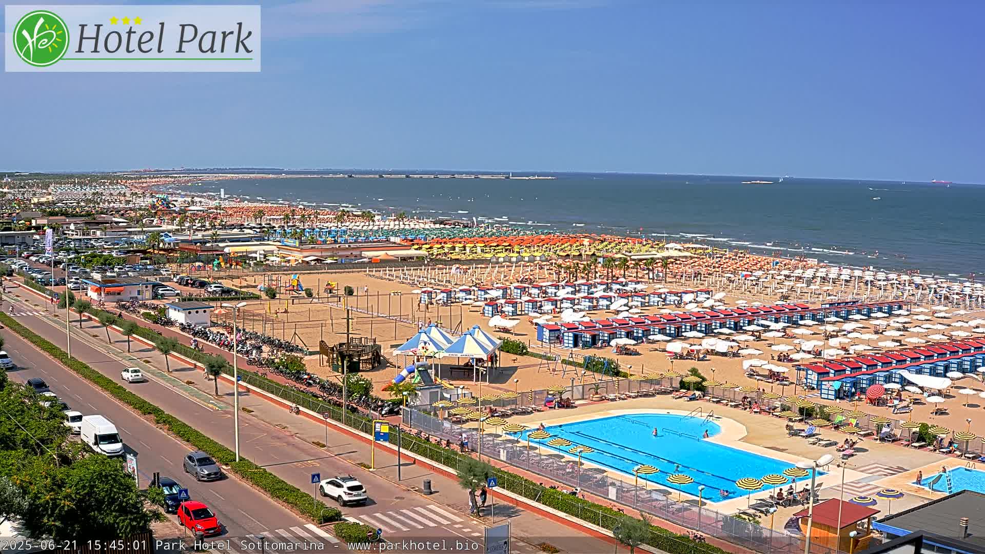 A sunny day at a beach resort shows a wide sandy beach with numerous umbrellas and beach cabanas, a swimming pool, and a road with cars.
