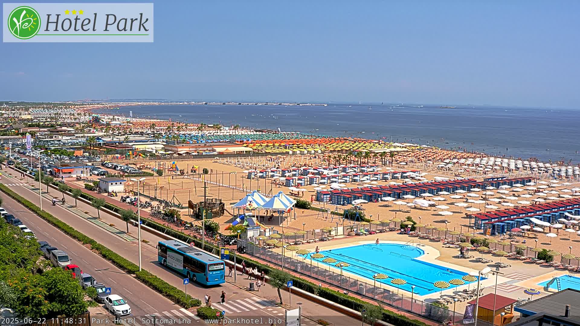 A beach scene on a sunny day shows a long stretch of sand with numerous beach umbrellas and cabanas, a swimming pool, and a road with cars and a bus.