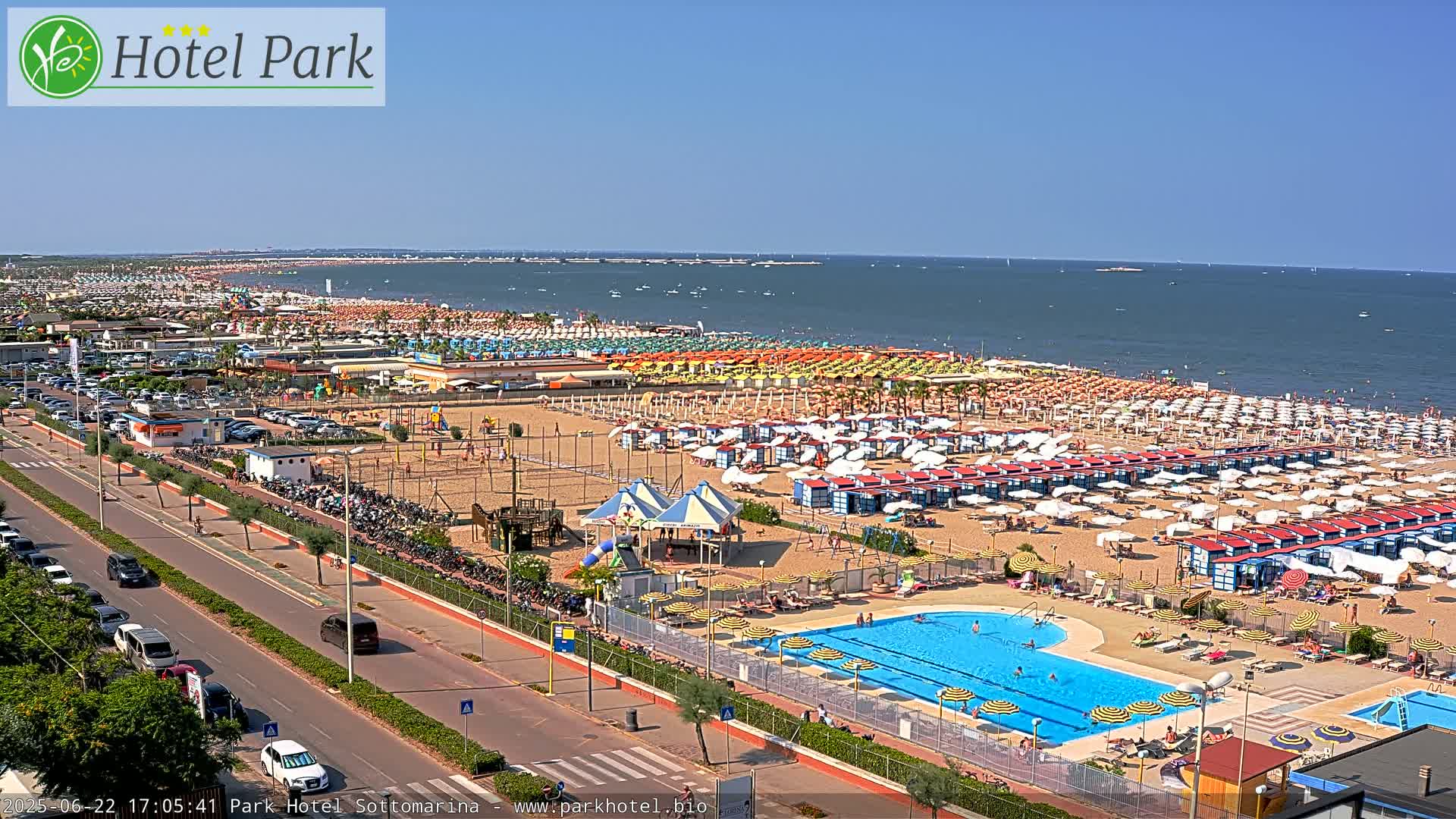 A sunny day at a beach resort shows a wide sandy beach filled with colorful umbrellas and beach cabanas, a swimming pool, and a road lined with parked cars.