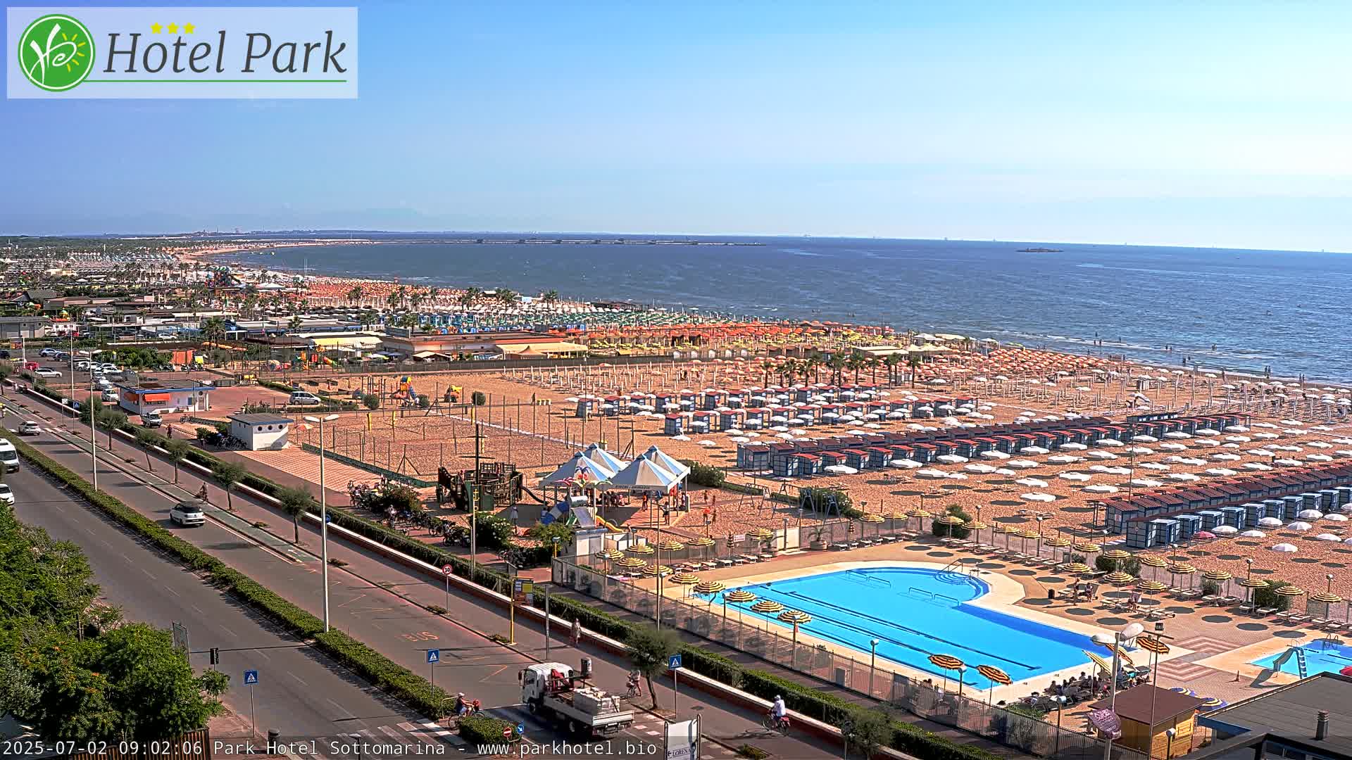 A sunny day at a beach resort shows a sandy beach lined with rows of umbrellas and beach chairs, a swimming pool, playground equipment, and a road with cars.