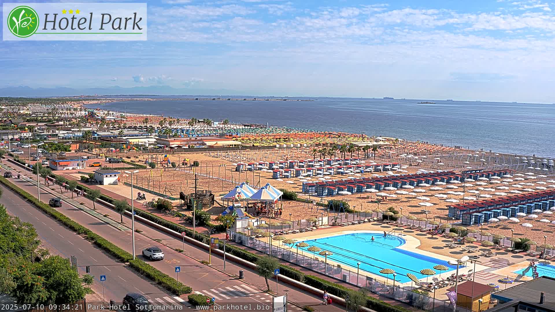 A sunny beach scene shows a large swimming pool, numerous beach umbrellas, and a playground next to a roadway with cars, all overlooking a calm ocean under a mostly clear sky.
