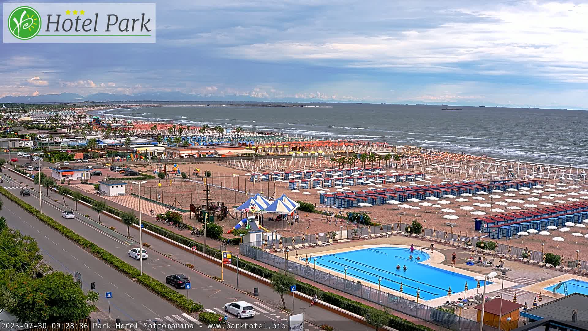 A sandy beach with numerous umbrellas and beach cabanas, a swimming pool, and a playground is shown, overlooking a calm sea under a partly cloudy sky.