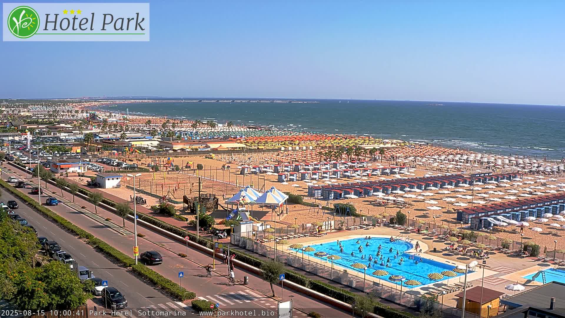 A sunny day at a beach resort shows a crowded sandy beach with numerous umbrellas and beach cabanas, a swimming pool with people in it, a road with cars, and a playground.