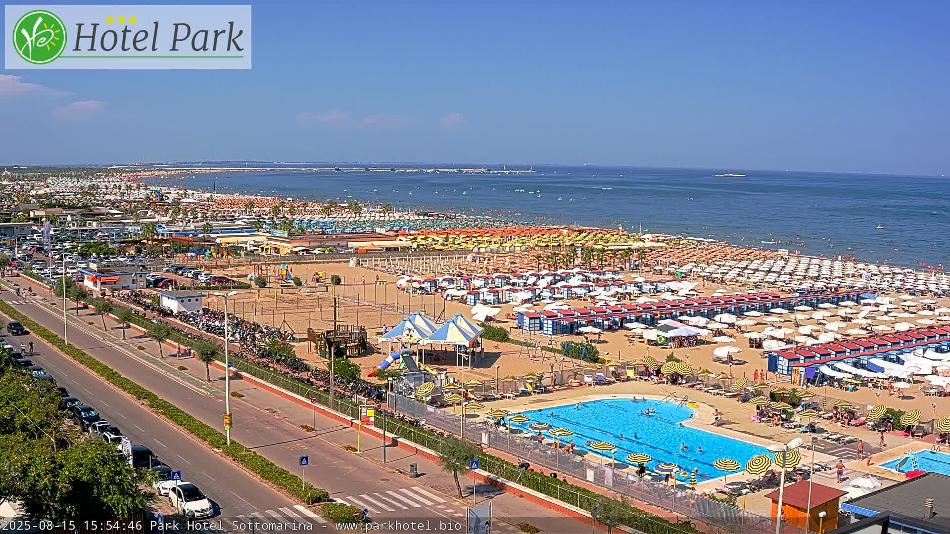 A sunny day at a beach resort shows a crowded sandy beach with numerous umbrellas and beach cabanas, a swimming pool with people in it, a road with cars, and a playground.