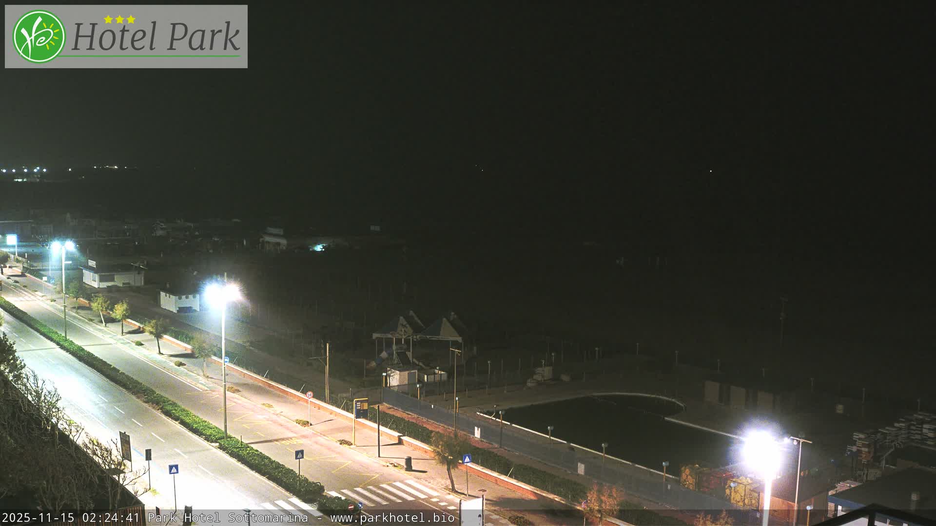 The image captures an outdoor night scene of a well-lit road and adjacent buildings bordering a dark expanse, likely a beach or open land, under a clear dark sky.