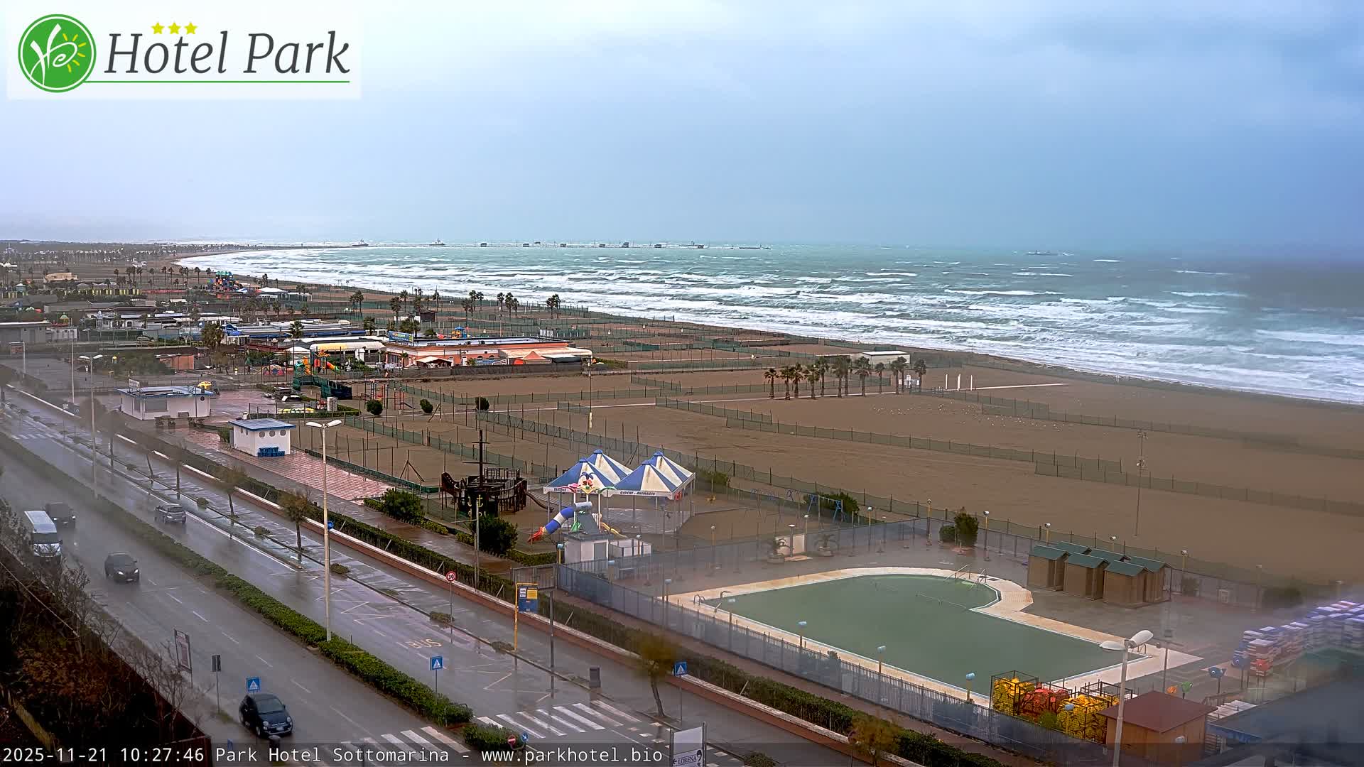 A wide aerial view captures a long, fenced beach with palm trees, an adjacent road with moving cars, a murky outdoor pool, and a choppy sea under an overcast sky, suggesting a rainy and windy day.