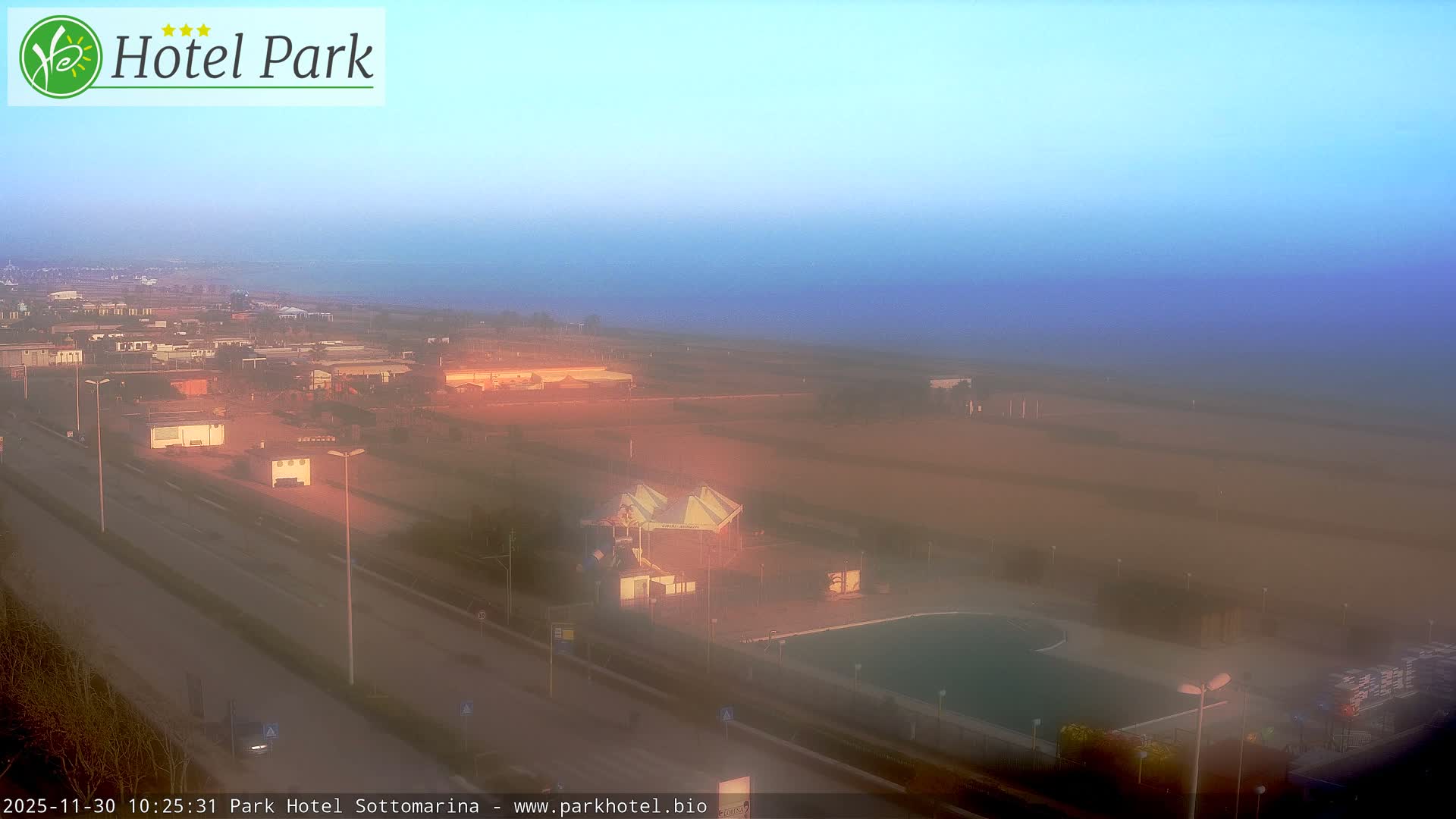 A hazy outdoor scene depicts a coastal town with a road, buildings, and a swimming pool complex in the foreground, leading to a distant, obscured ocean under a graduated blue sky.