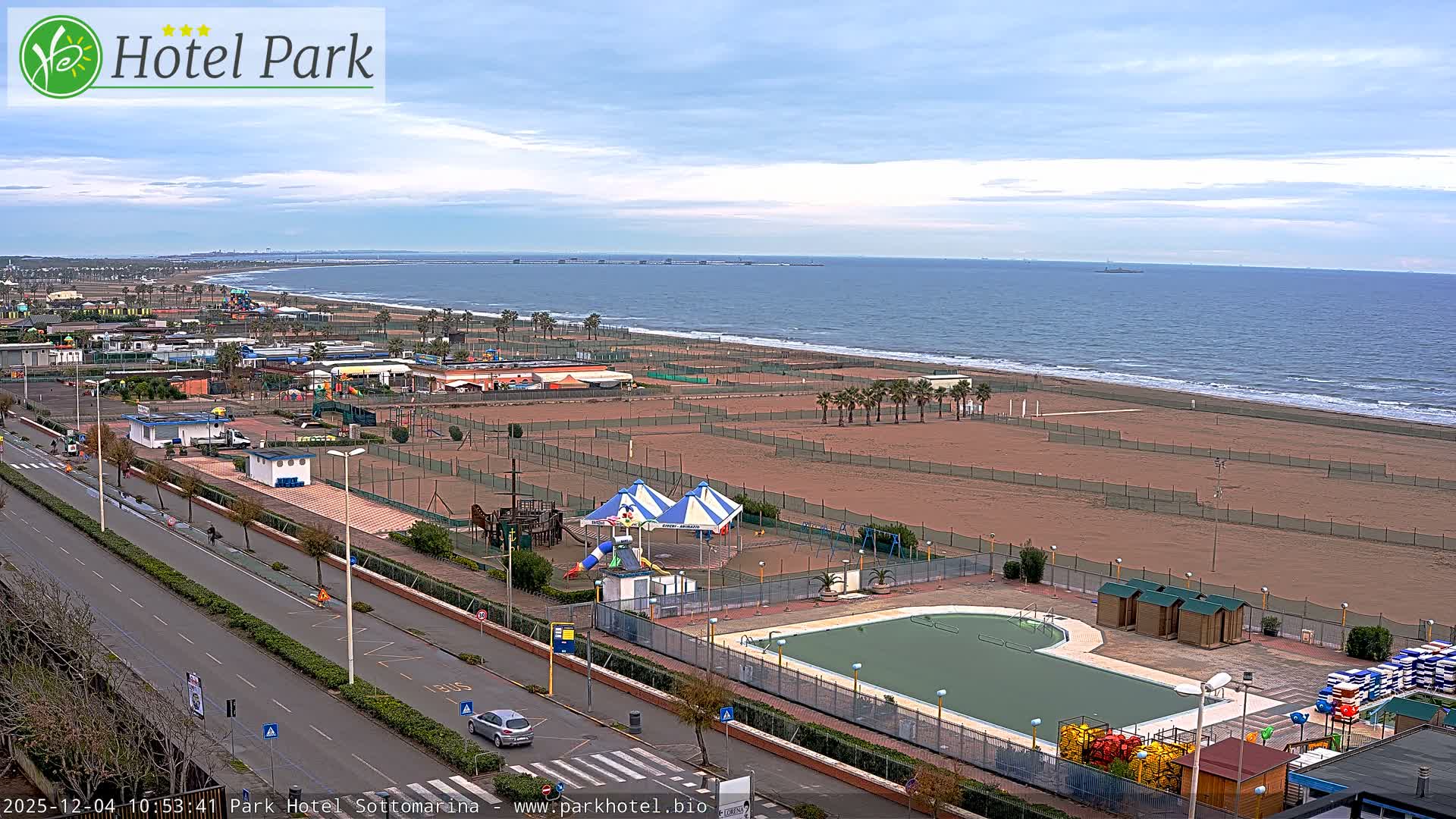 On an overcast day, a coastal resort town is seen from above, showcasing a multi-lane road, fenced sandy beaches with recreational areas and an outdoor swimming pool, all bordering a calm sea with a distant pier.