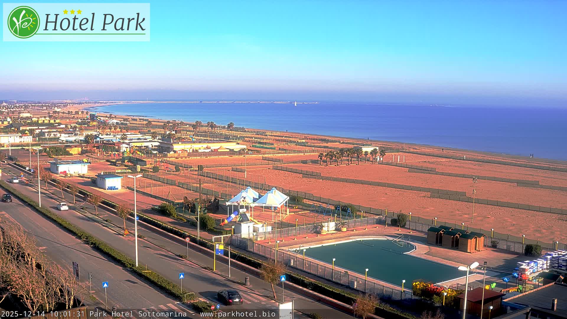 On an overcast day, a coastal resort town is seen from above, showcasing a multi-lane road, fenced sandy beaches with recreational areas and an outdoor swimming pool, all bordering a calm sea with a distant pier.