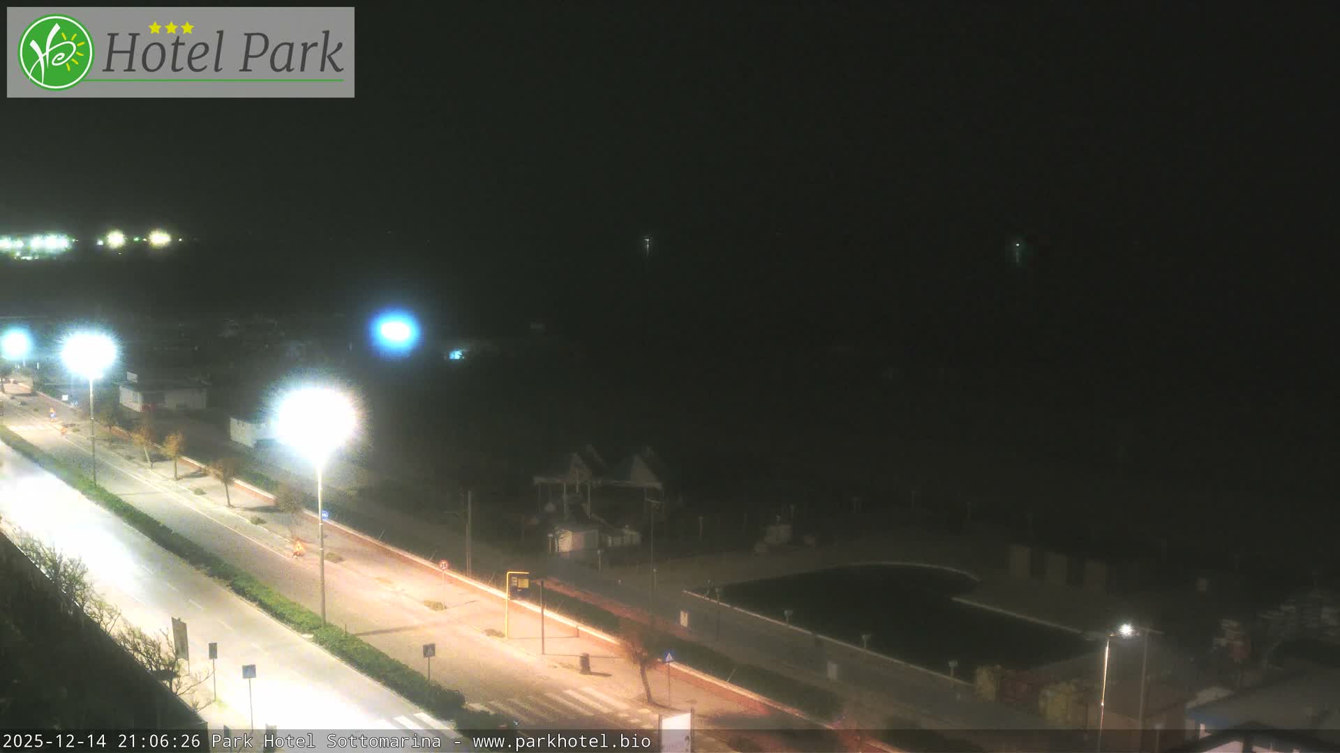 On an overcast day, a coastal resort town is seen from above, showcasing a multi-lane road, fenced sandy beaches with recreational areas and an outdoor swimming pool, all bordering a calm sea with a distant pier.