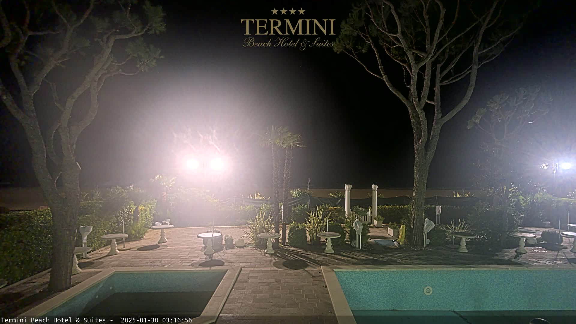 At night, a hazy, brightly lit patio area with two swimming pools is surrounded by trees and landscaping.