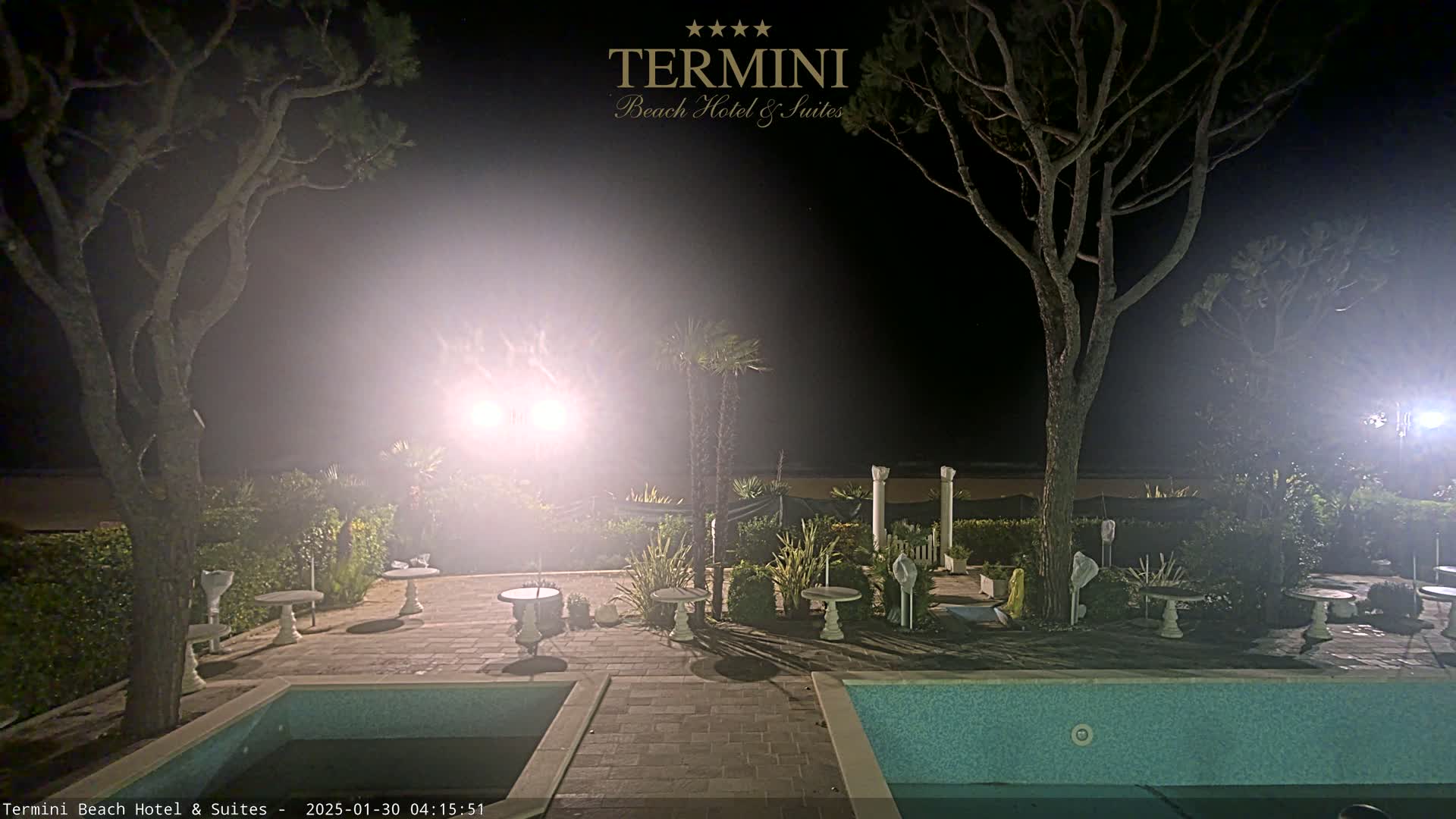 A nighttime view of a hotel patio area with a swimming pool, trees, and small tables, illuminated by bright lights under a hazy sky.