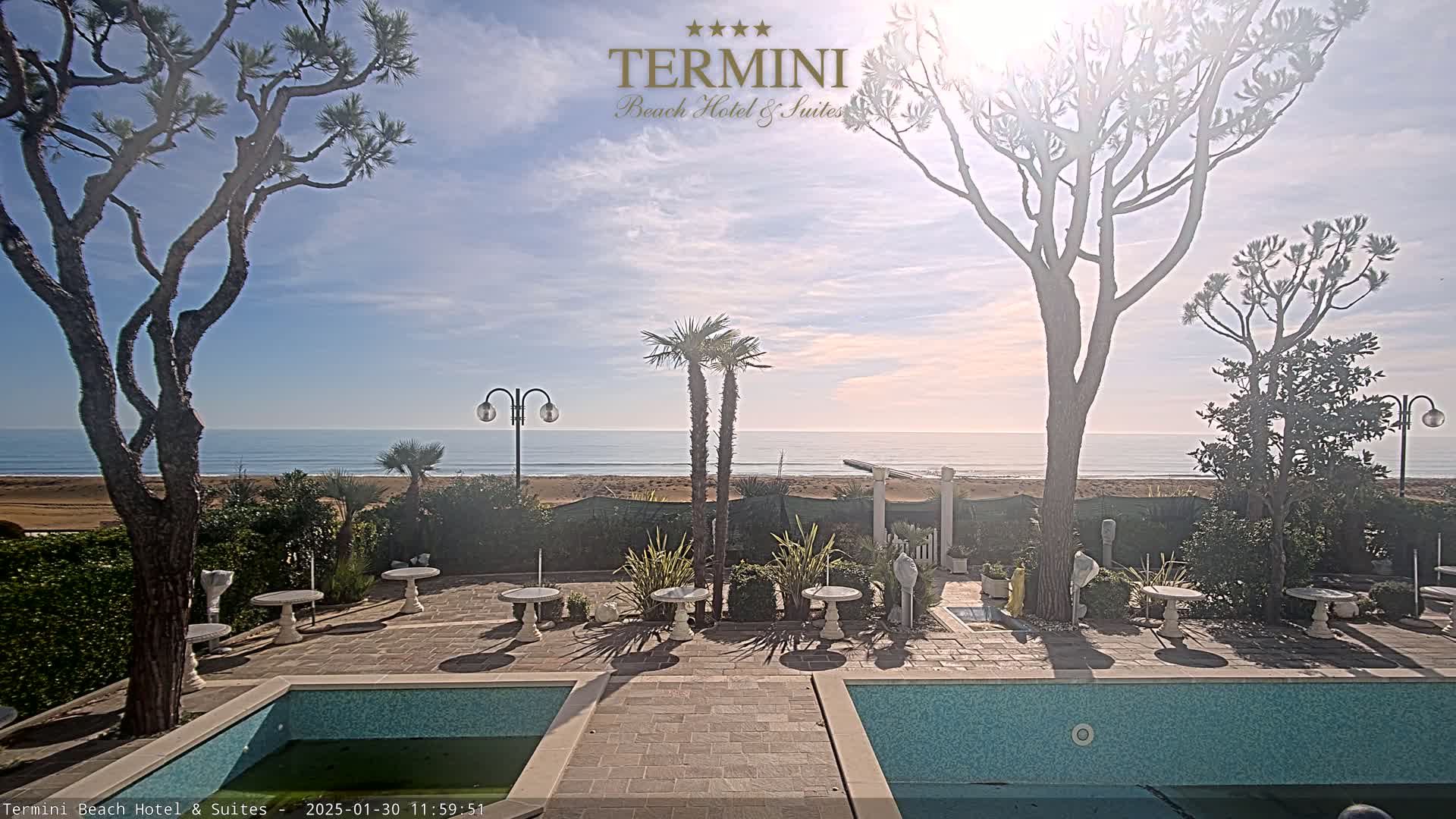 A patio with a swimming pool overlooks a calm ocean under a mostly sunny sky.