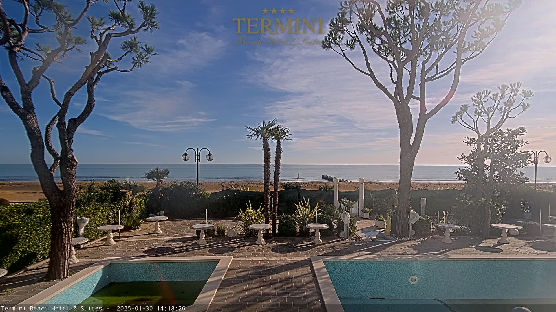 A tranquil outdoor scene with a clear blue sky, a calm ocean, a sandy beach, and a patio area with two swimming pools and small tables, surrounded by trees and landscaping.