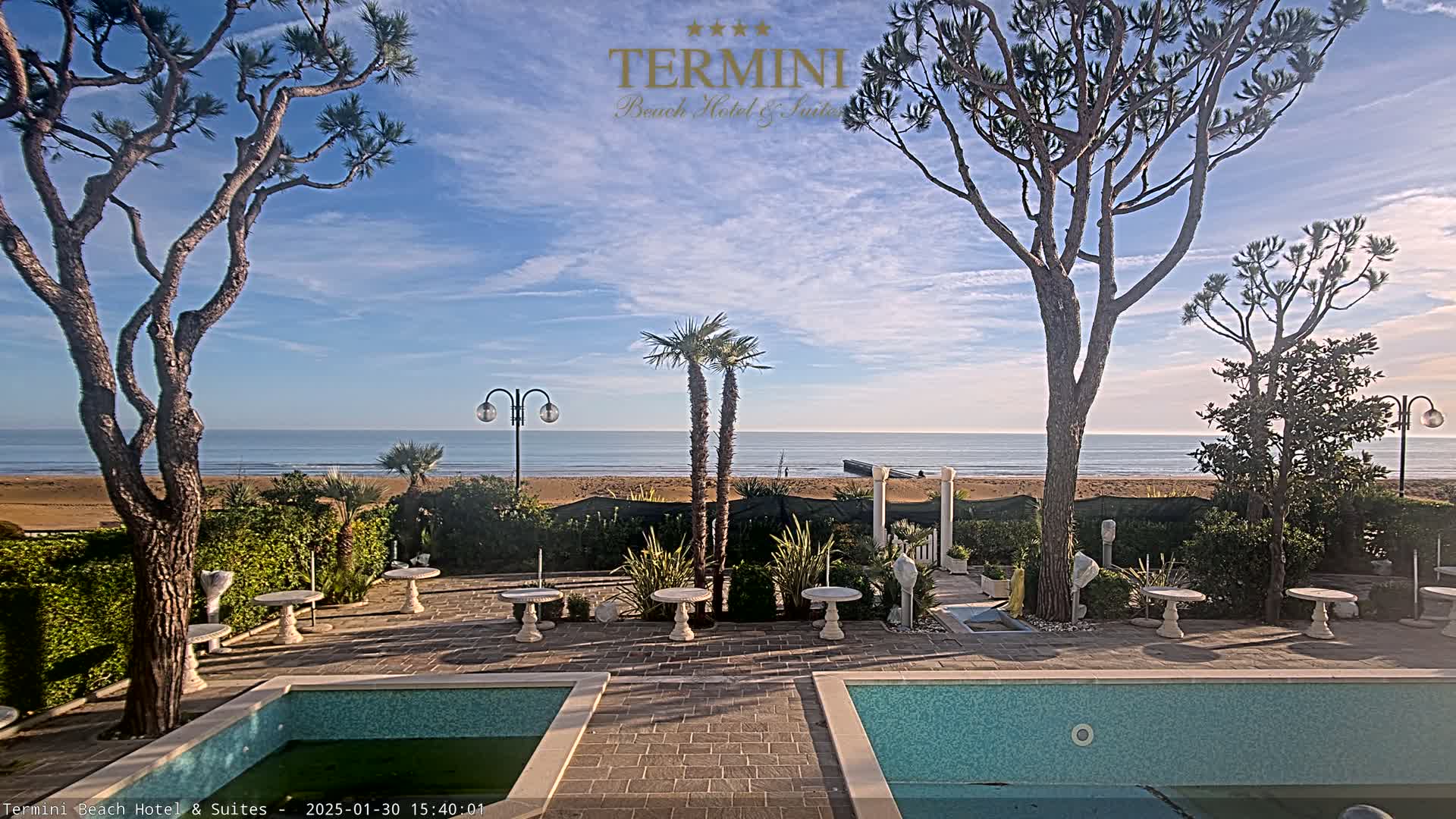 A patio with two small pools overlooks a calm ocean beach under a mostly sunny sky.