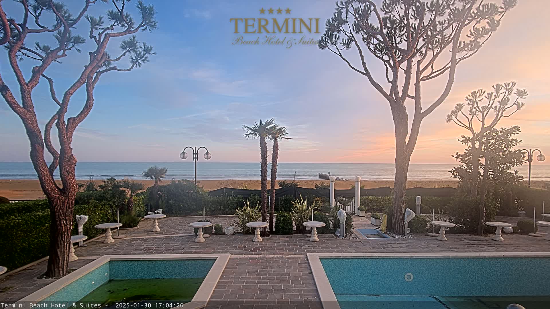 A tranquil outdoor patio scene with two pools overlooks a calm ocean beach at sunset.