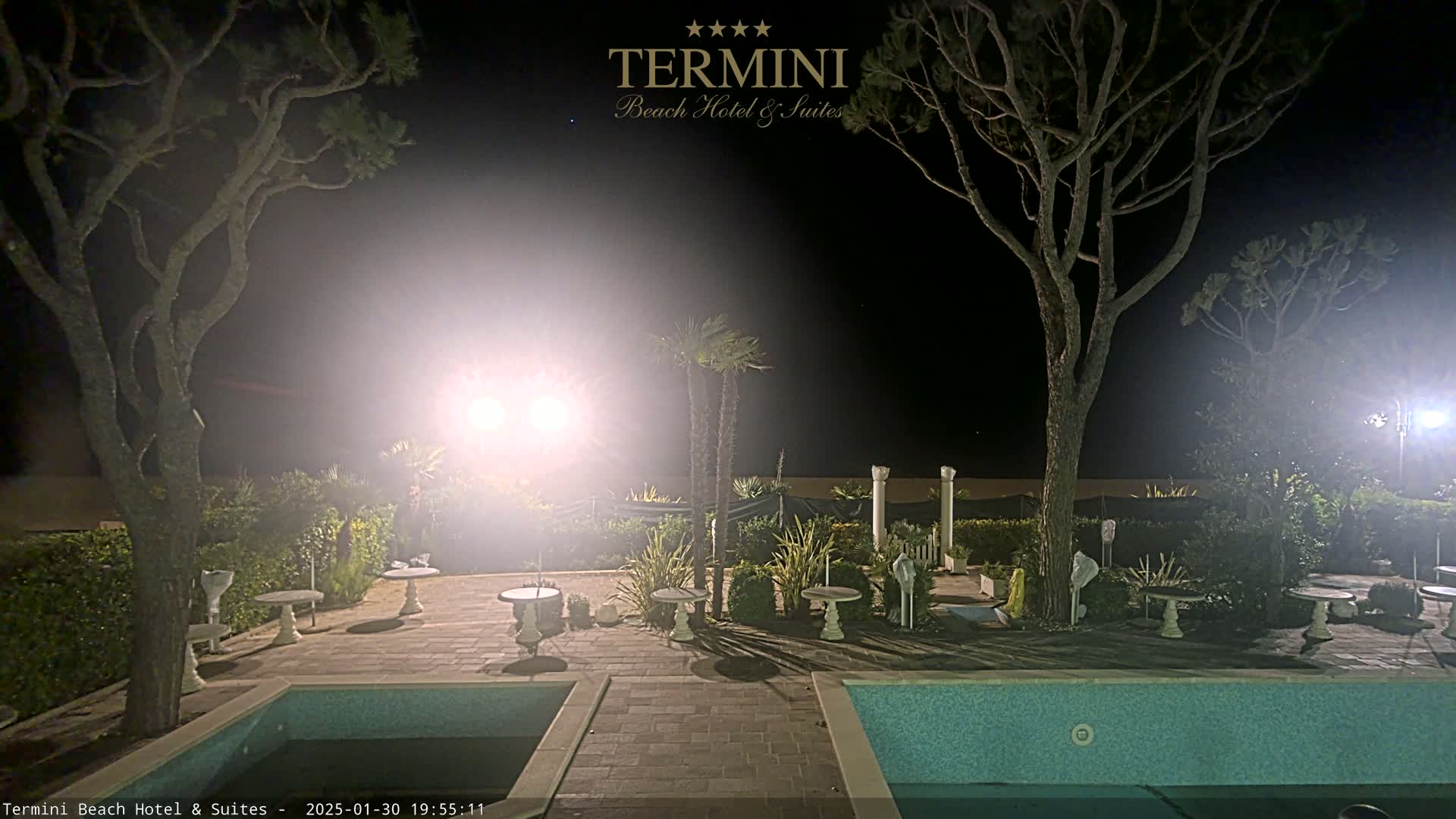 A nighttime view of a hotel courtyard with two swimming pools, several small tables, palm trees, and pine trees illuminated by bright lights.