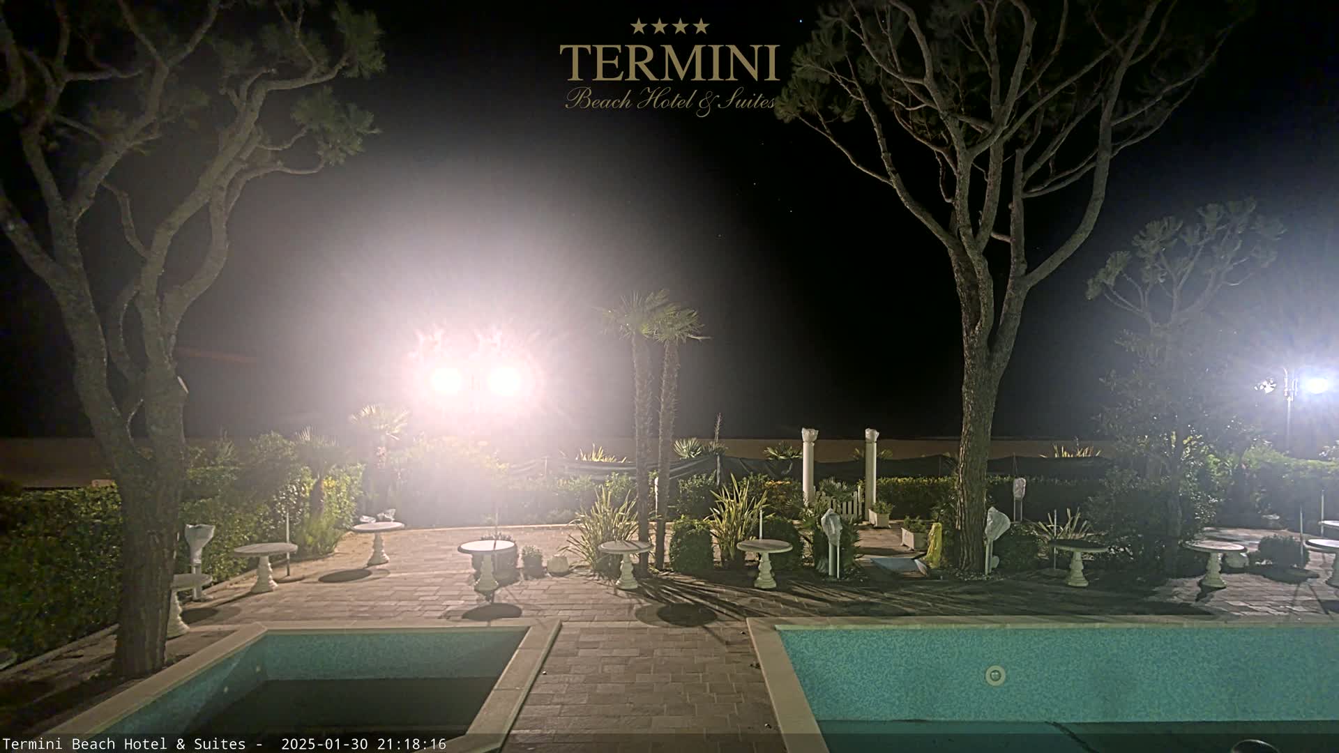 A nighttime view of a pool area with two trees, several small tables, and palm trees, illuminated by bright lights.