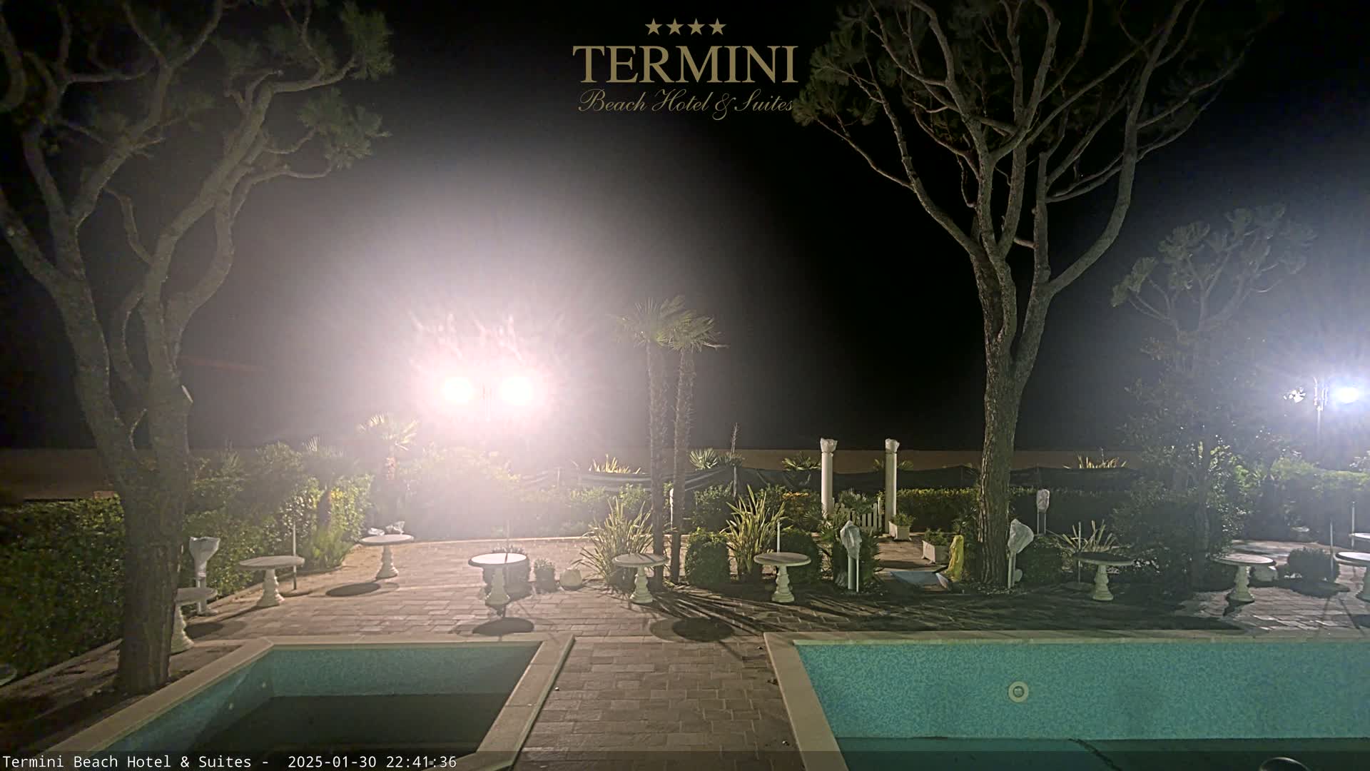 A nighttime view of a pool area with surrounding landscaping, trees, and patio furniture, illuminated by bright lights.