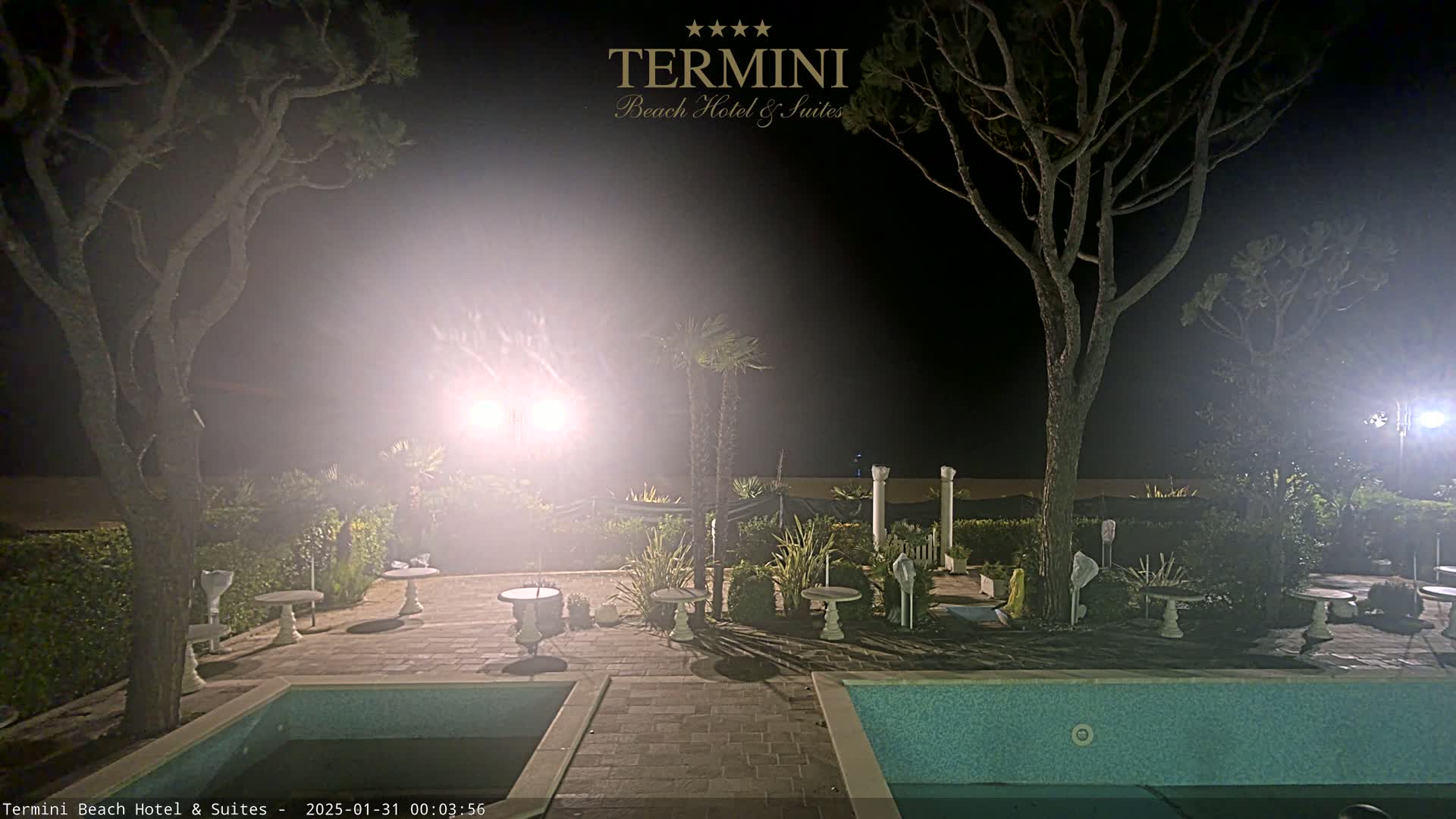 A hazy, moonless night scene shows a patio area with two swimming pools, small tables, and several trees illuminated by bright lights.