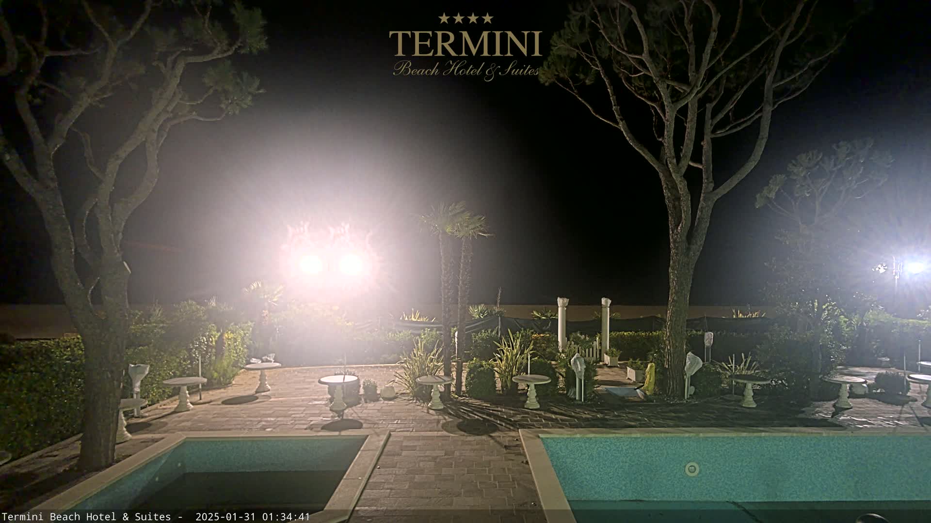 A nighttime view of a resort area with a pool, patio furniture, and trees illuminated by bright lights.