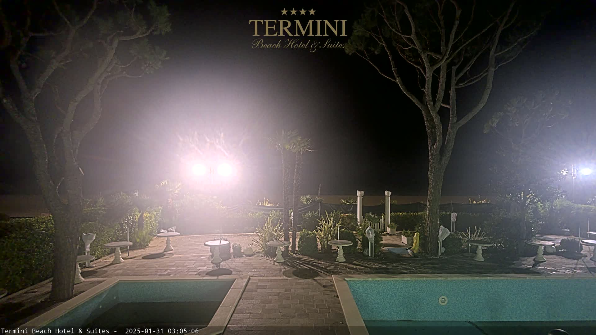 A hazy night scene shows an outdoor area with two swimming pools, patio furniture, and trees illuminated by bright lights.