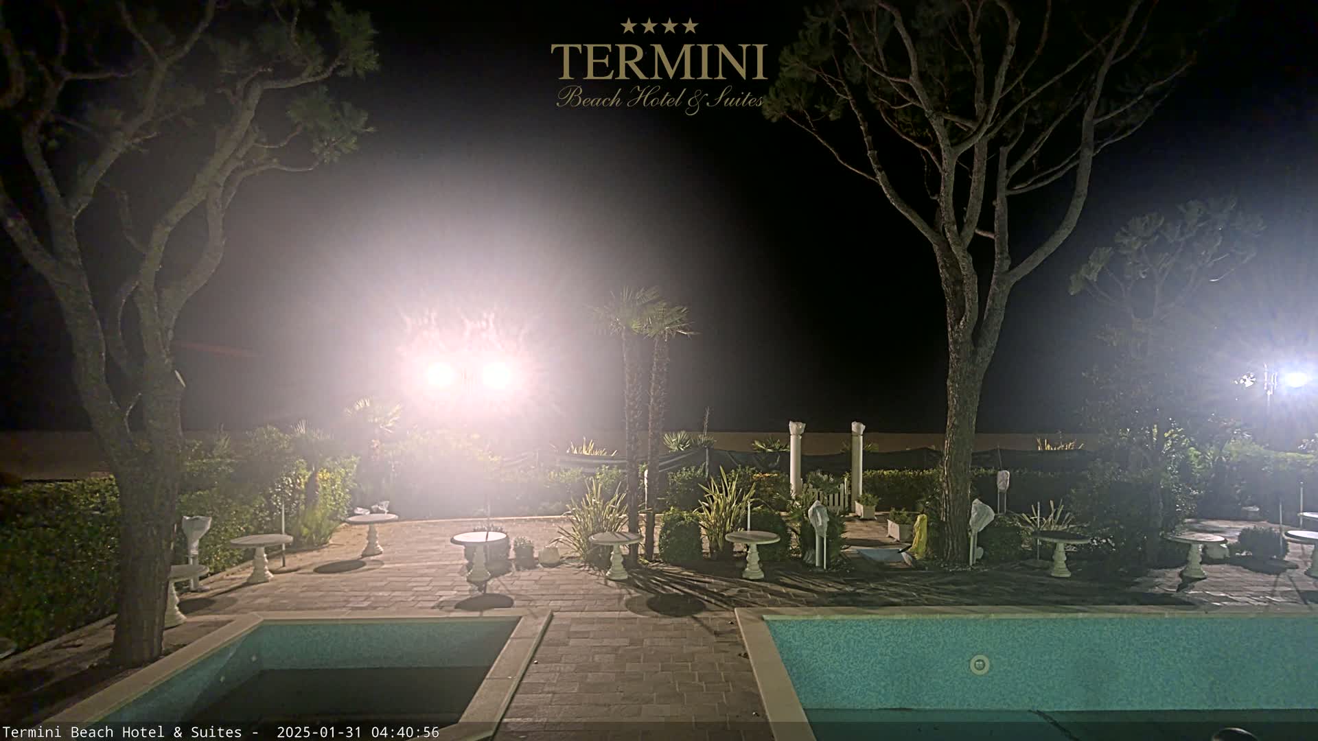 A nighttime view of a hotel patio with two pools, several small tables, and trees, illuminated by bright lights in hazy conditions.