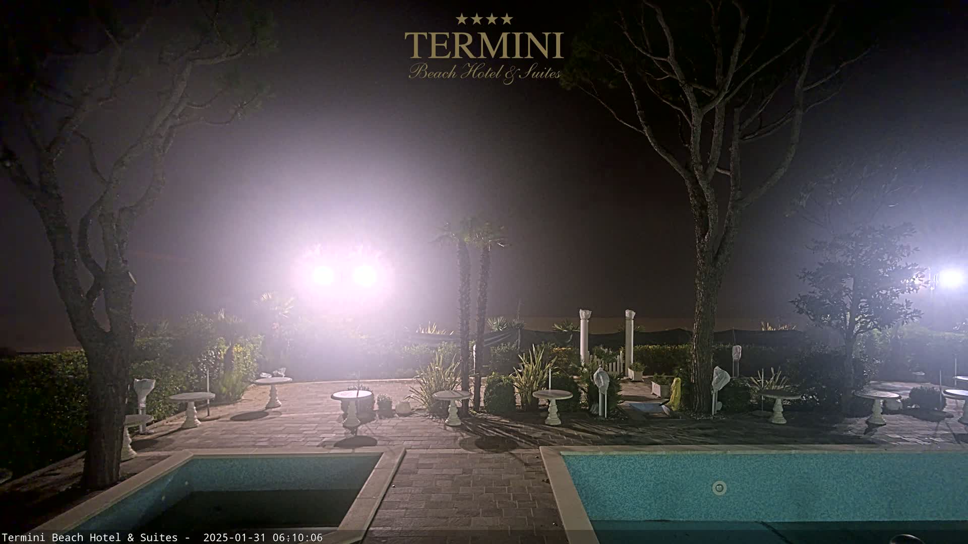 A fog-shrouded nighttime scene shows a patio area with a pool, small tables, and trees, illuminated by bright lights.