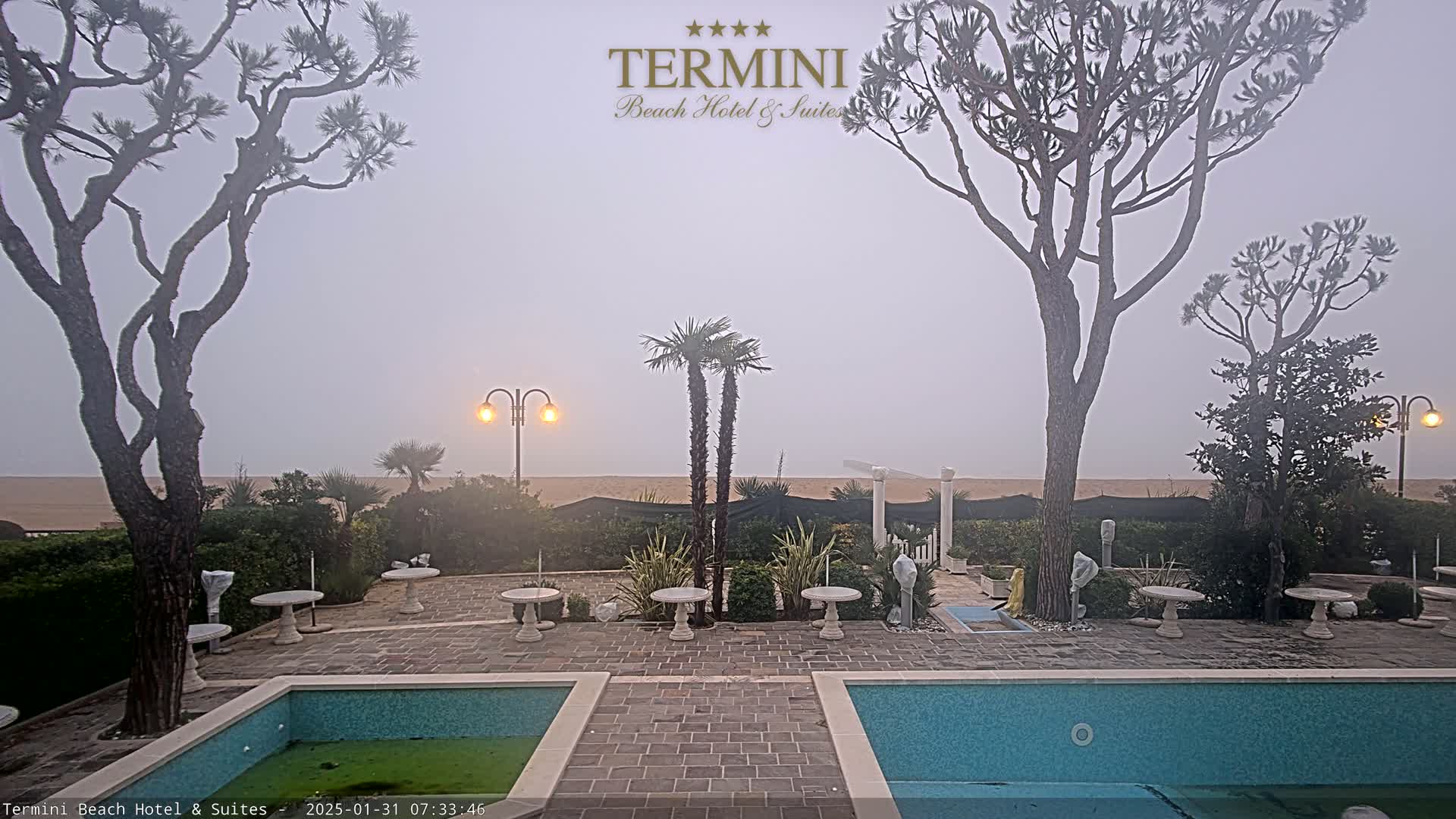 A foggy beach scene is visible from a patio with two swimming pools and small tables, surrounded by trees and landscaping.