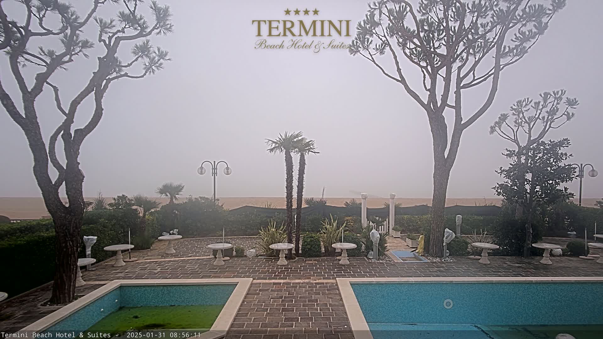A foggy beach scene is visible beyond a courtyard with two swimming pools, stone tables, and several trees.