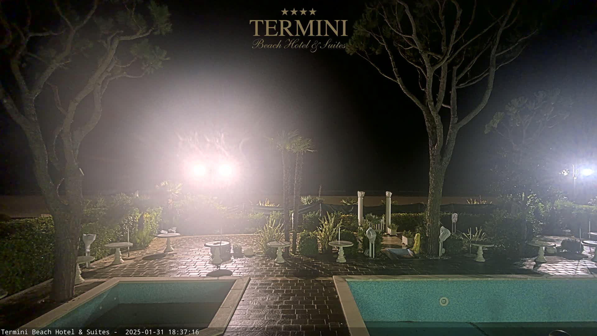 A nighttime view of a patio area with two swimming pools, surrounded by trees and plants, under hazy, brightly lit conditions.