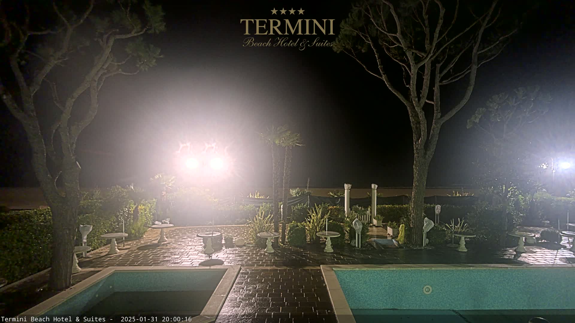 At night, a courtyard with two swimming pools is illuminated by bright lights, surrounded by trees and shrubs.