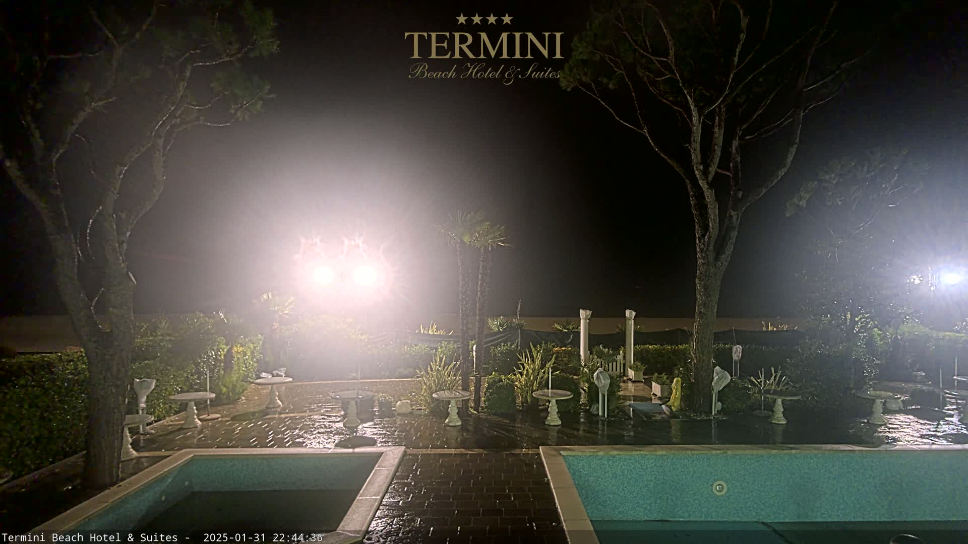At night, under a misty sky, an outdoor patio area with two pools and several small tables is illuminated by bright lights.