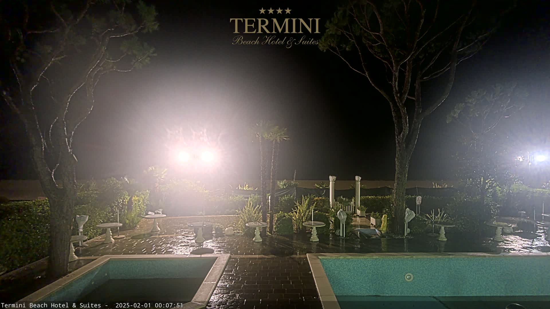 A nighttime scene shows a wet patio area with two pools and several small tables, surrounded by trees and shrubs under hazy, bright lights.