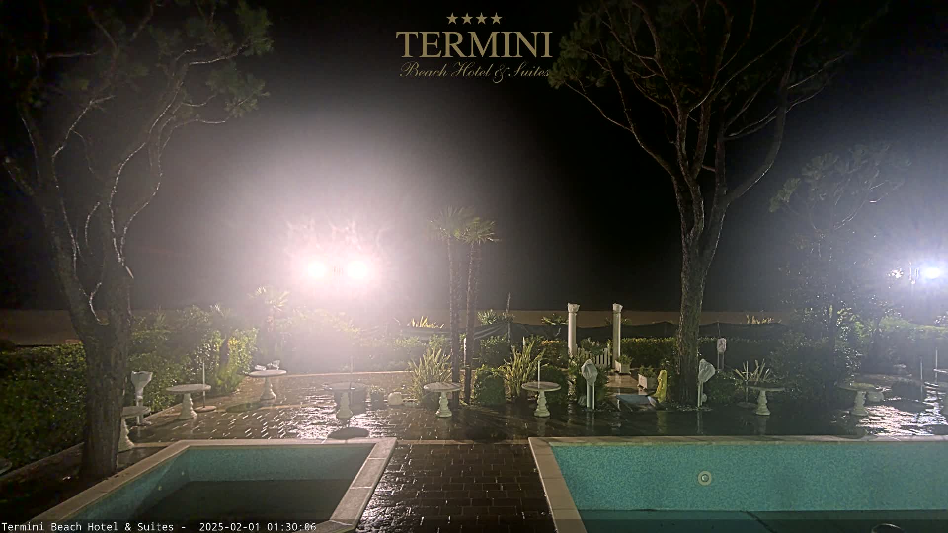 A nighttime view of a resort area with two pools, patio furniture, and trees under foggy conditions.