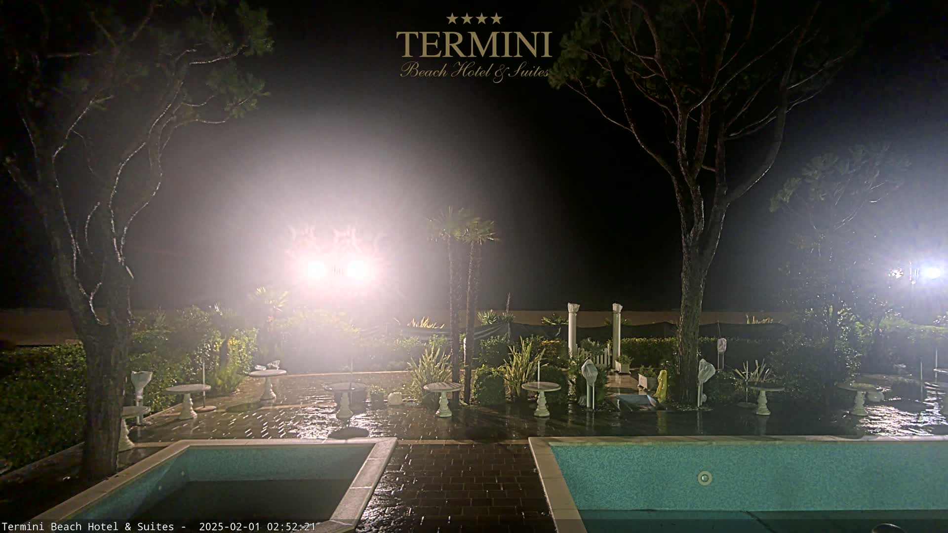 At night, under hazy light, a wet patio area with two pools and small tables is surrounded by trees and shrubs.