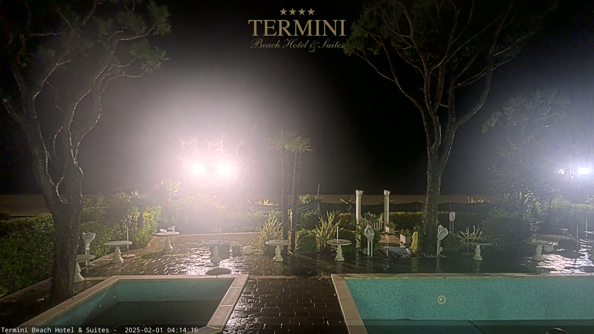 At night, under hazy lighting conditions, a wet patio area surrounds a pool, with trees and small tables visible in the background.