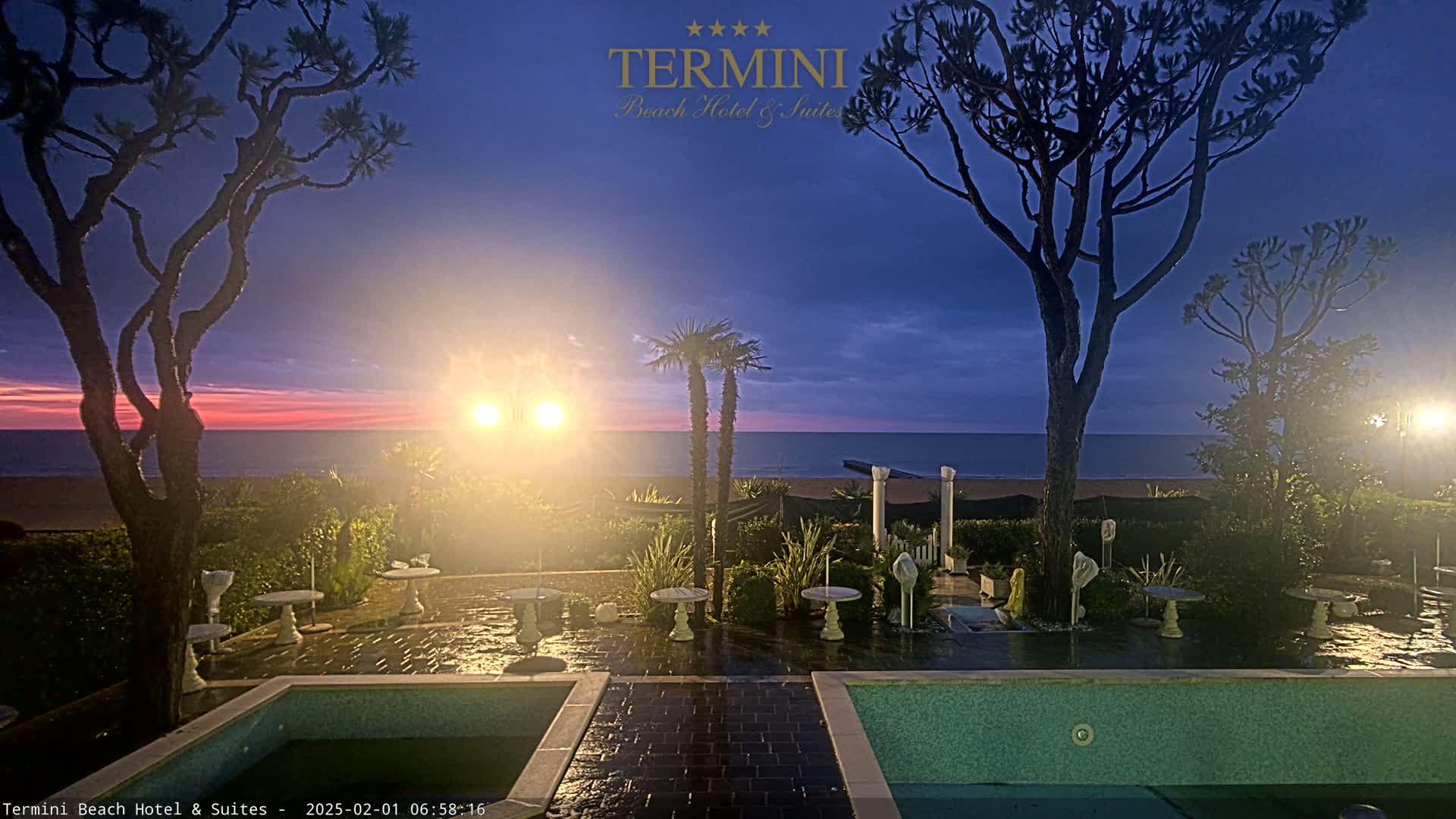 An outdoor patio scene at twilight shows a calm ocean in the background, with trees, plants, small tables, and a pool in the foreground.