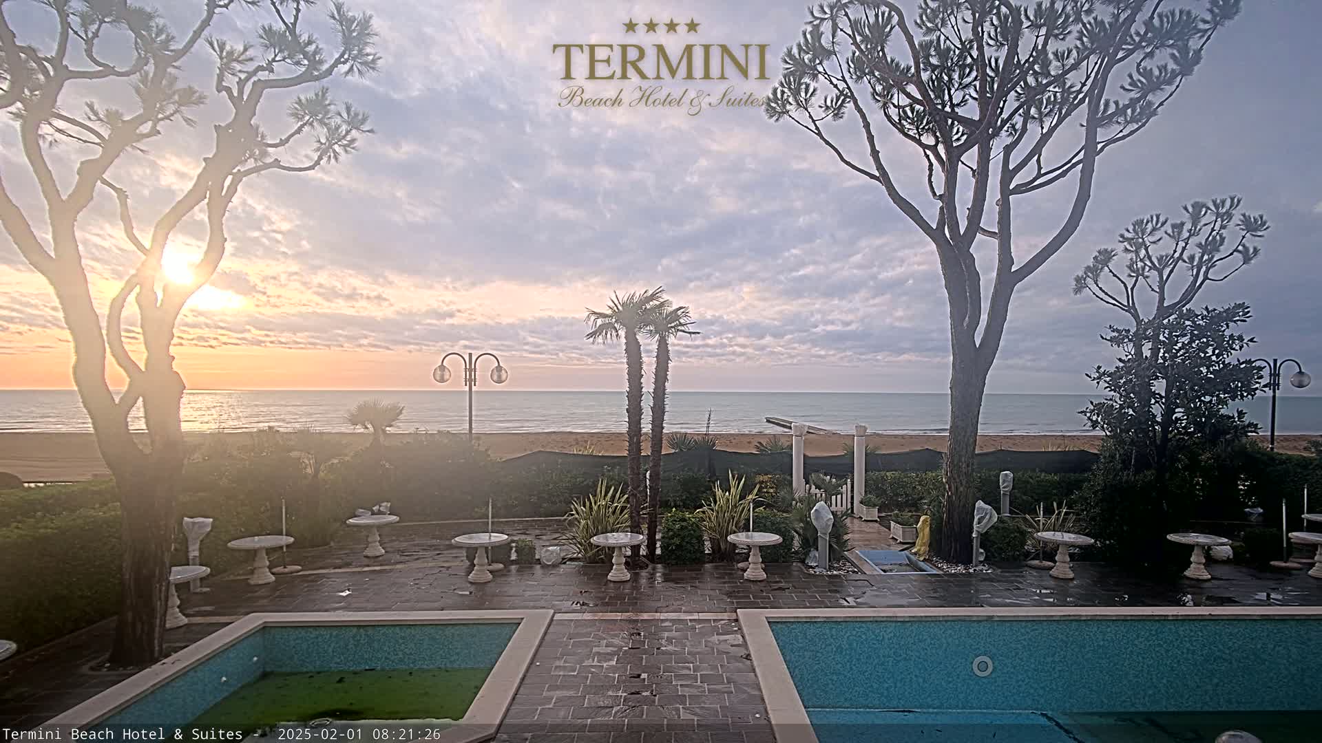 A tranquil sunrise over a calm ocean is visible from a patio with two swimming pools, several small tables, and tall trees.