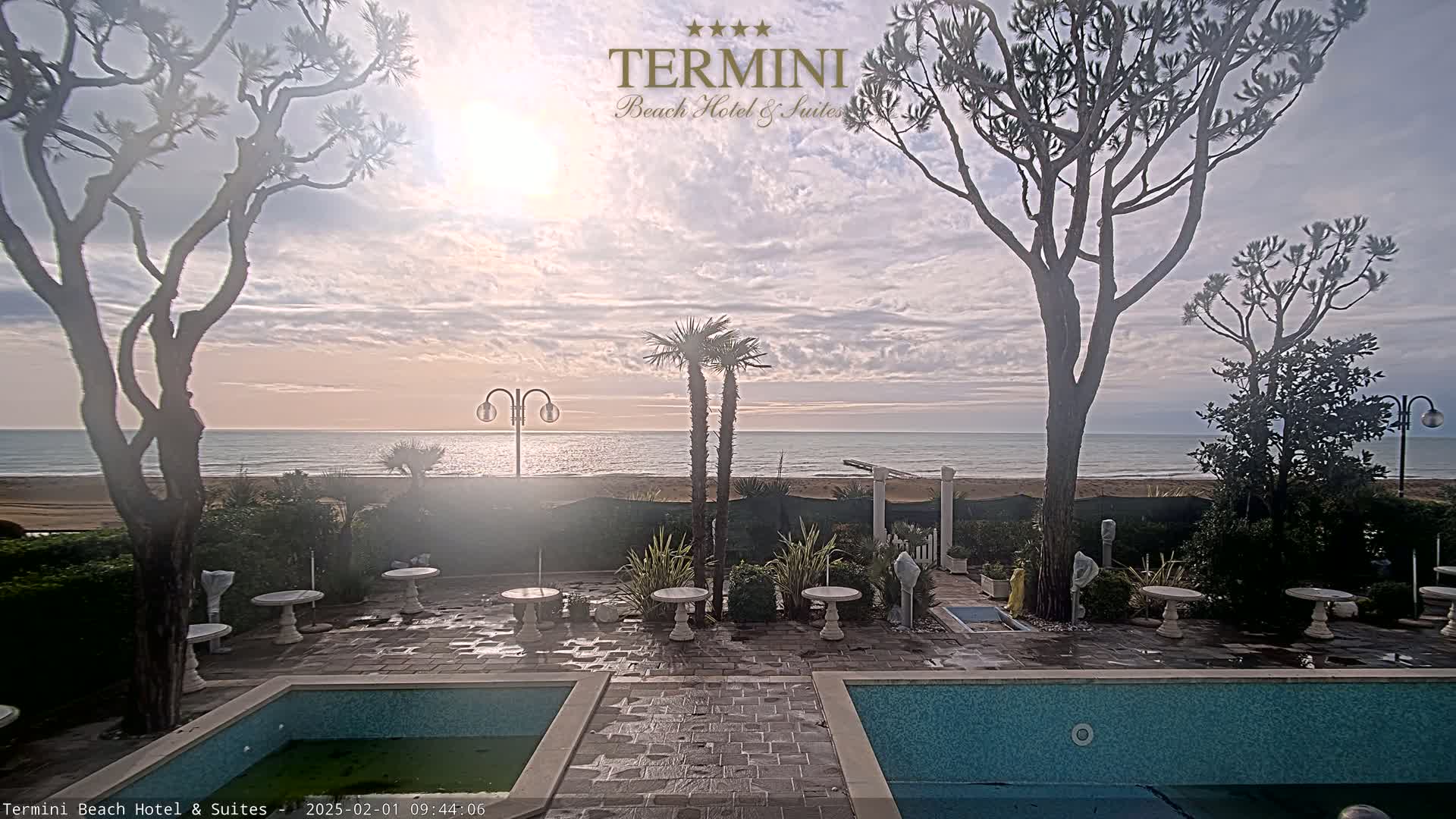 A tranquil beach scene at sunrise, viewed from a patio area with two pools, several small tables, and tall trees, under a partly cloudy sky.