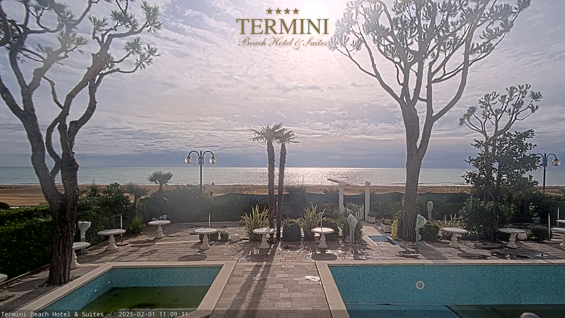 A partly cloudy day shows a tranquil ocean scene viewed from a patio with a swimming pool, featuring several trees and small tables.