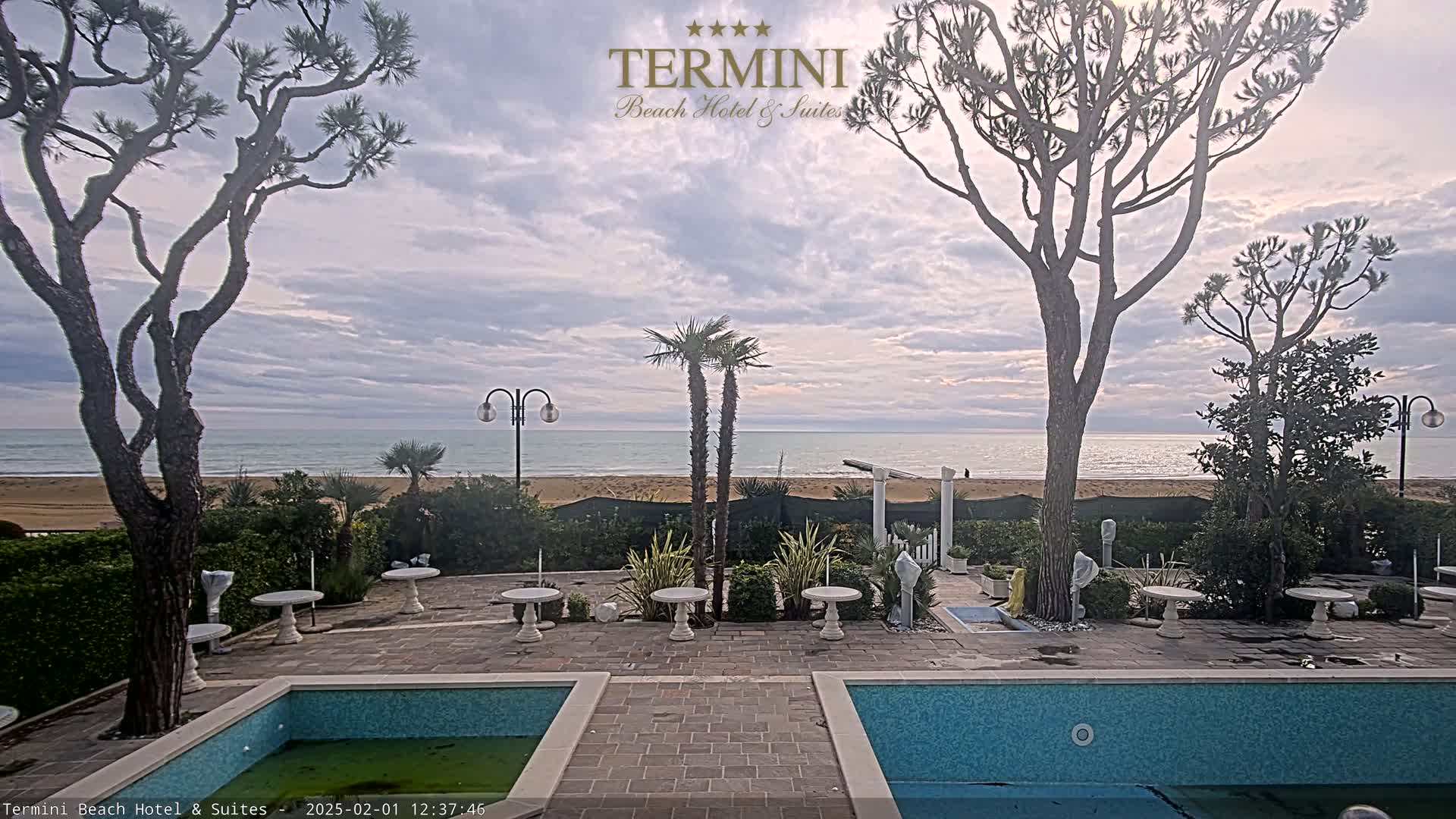 An outdoor patio area with two pools overlooks a calm ocean under a cloudy sky, featuring several trees and small tables scattered around.
