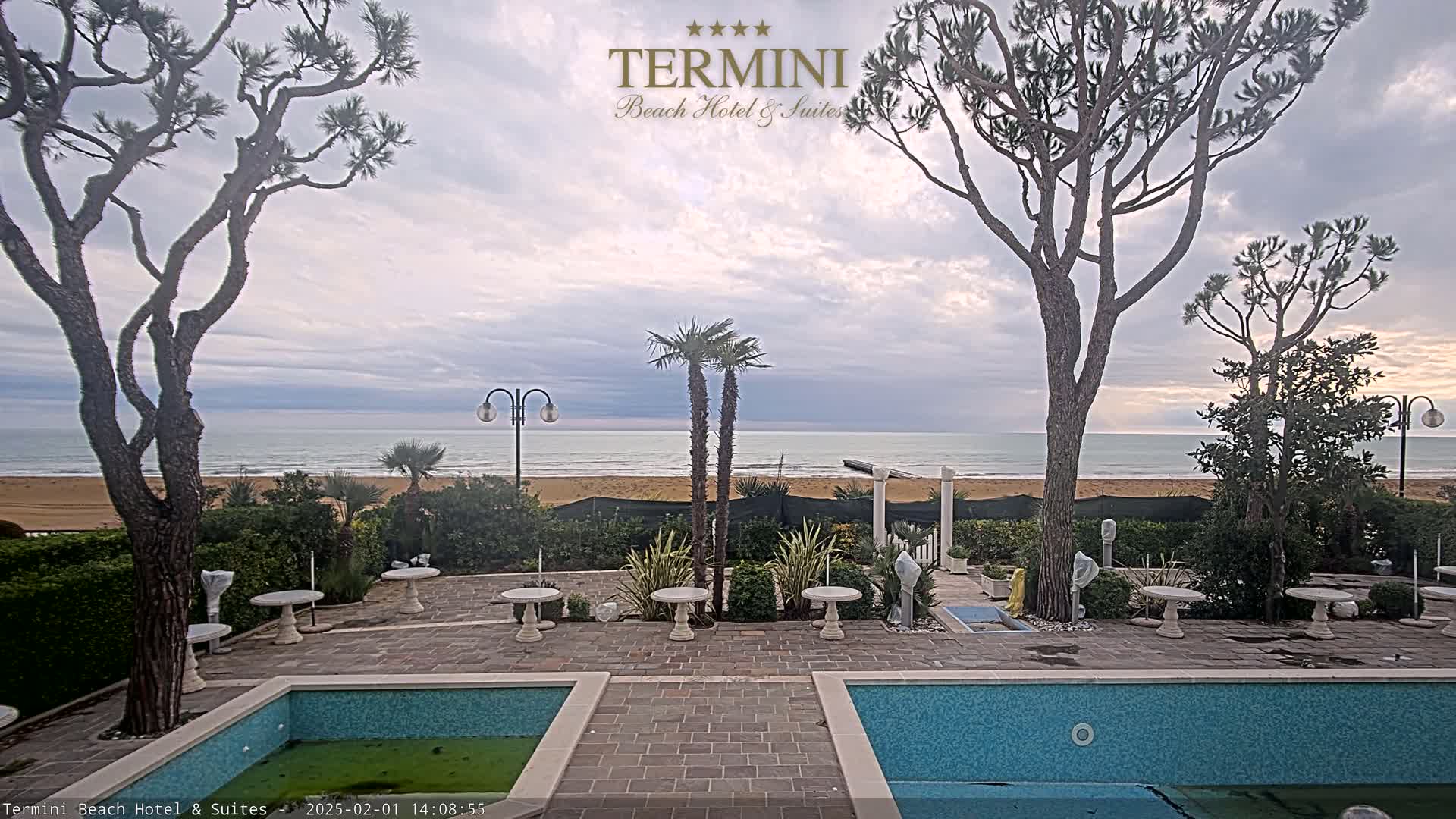 A patio area with two small swimming pools overlooks a sandy beach and calm ocean under a cloudy sky.