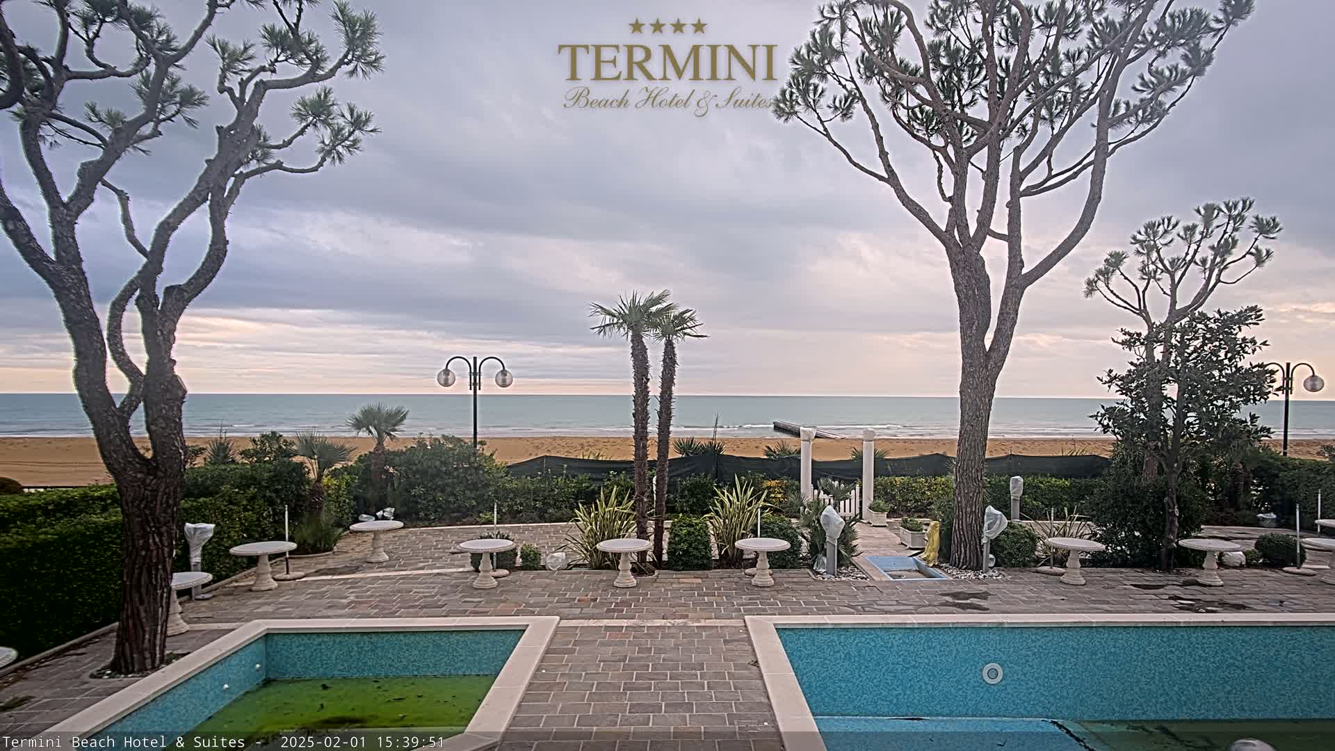 An outdoor patio area with two swimming pools overlooks a sandy beach and calm ocean under a cloudy sky.
