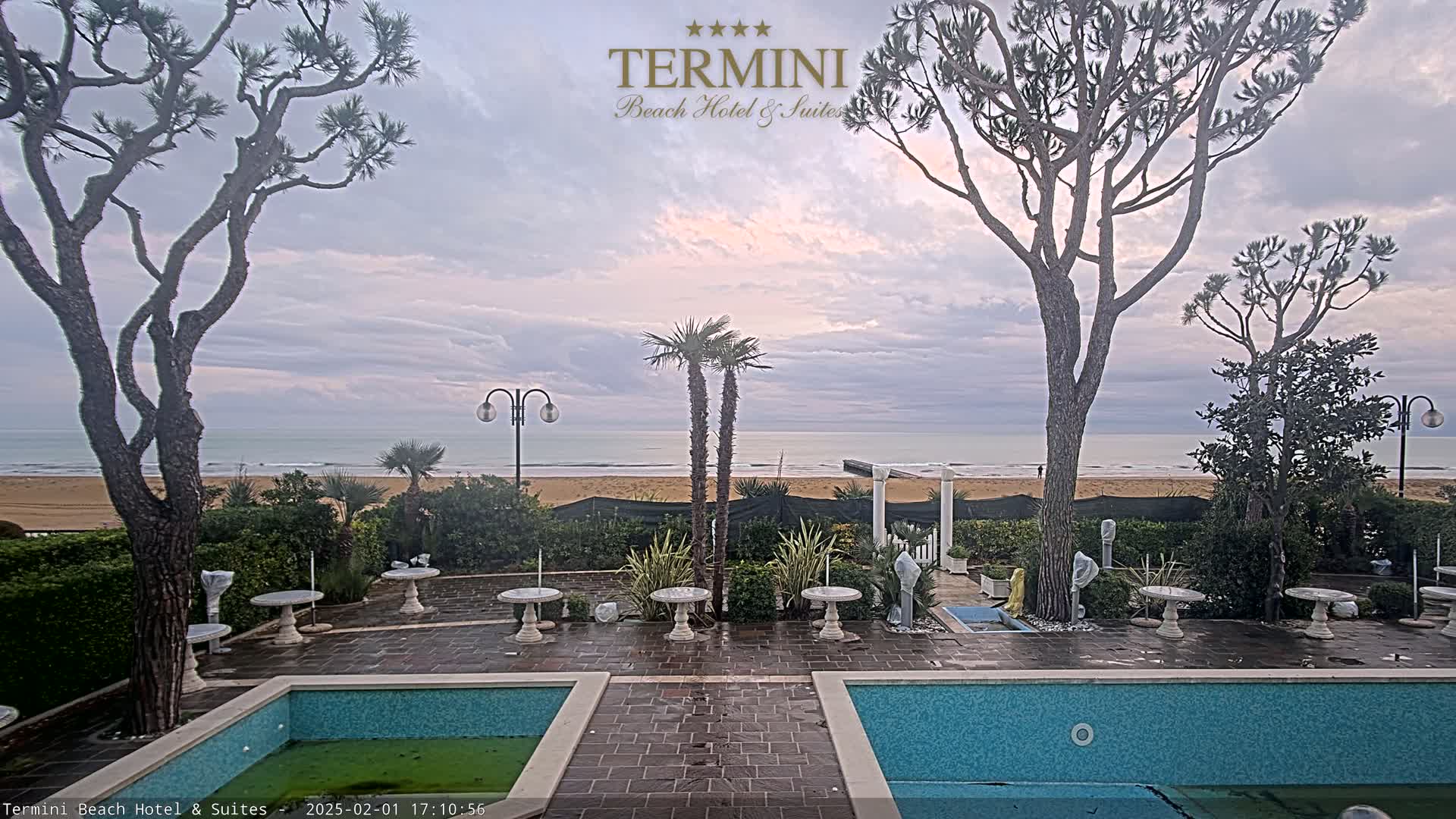 A patio with two swimming pools overlooks a calm ocean beach under a cloudy sky.