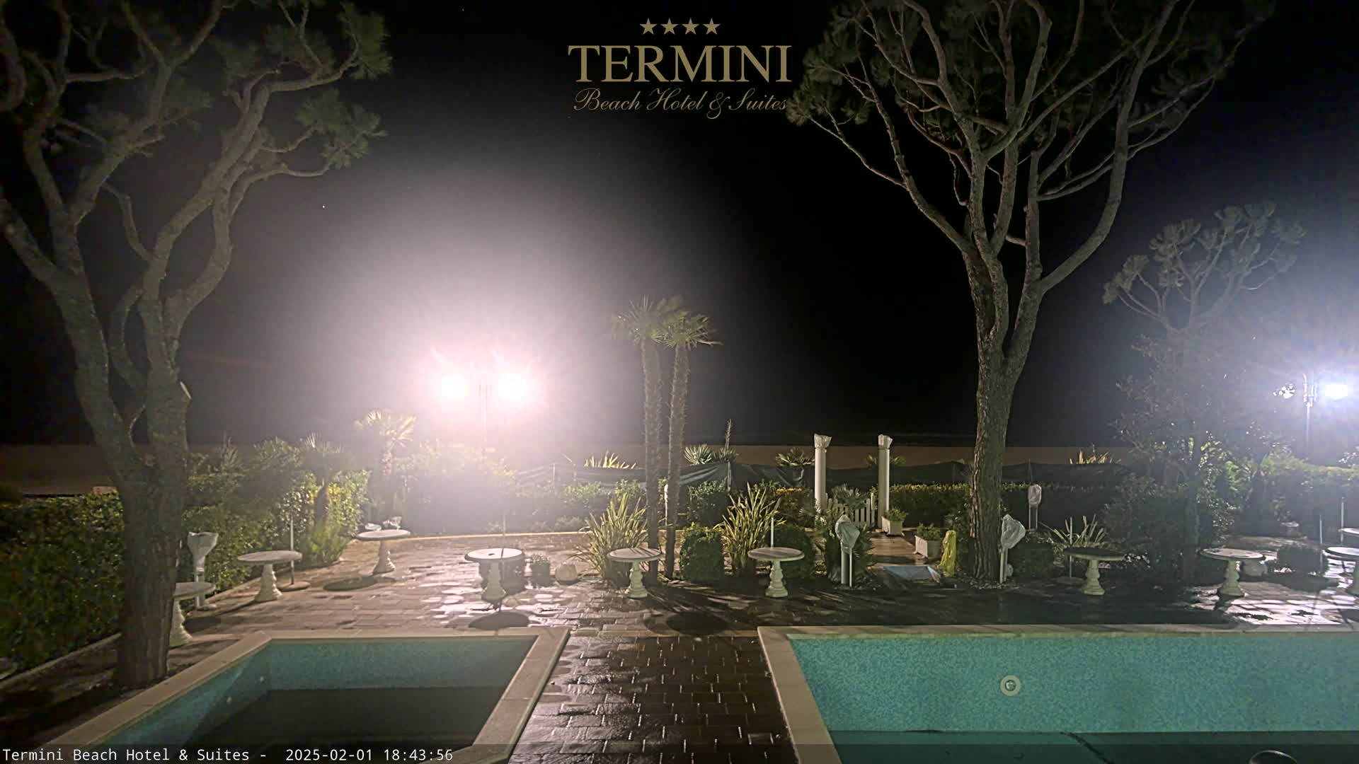 At night, a courtyard with two pools and several small tables is lit by bright lights under a hazy, dark sky.