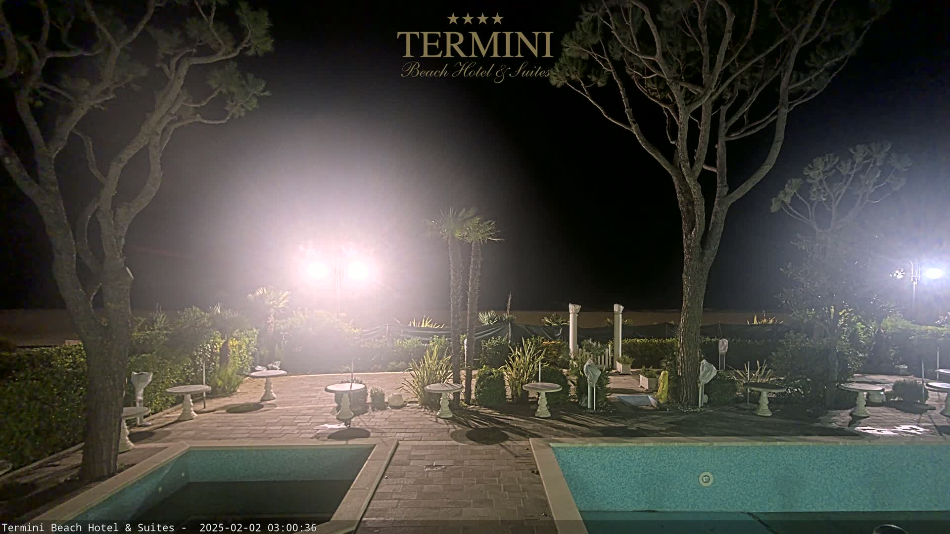 A nighttime view of a resort patio area with two pools, several small tables, and trees illuminated by bright lights.