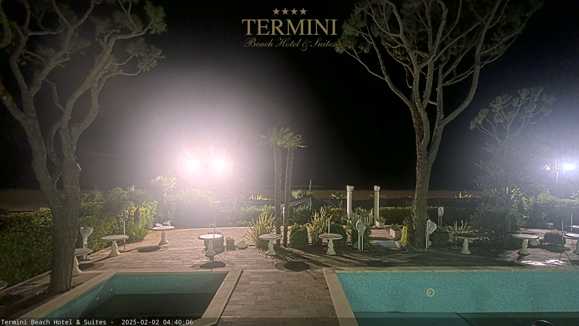 A nighttime view of an outdoor pool area with patio furniture, trees, and bright lights illuminating the scene.