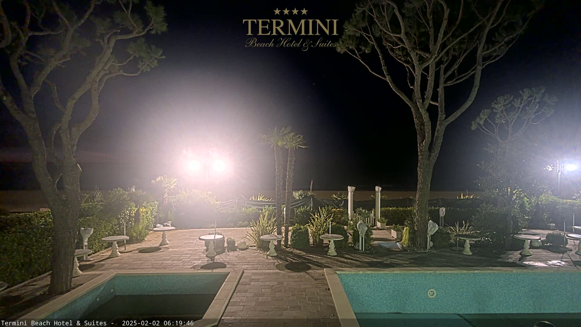 A nighttime view of a patio area with two swimming pools, several small tables, and palm trees, illuminated by bright lights.