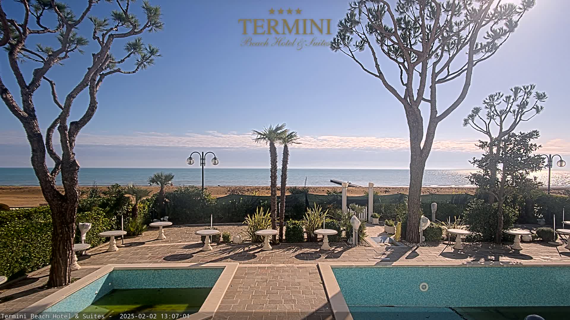 A patio with a swimming pool overlooks a calm ocean under a sunny sky.